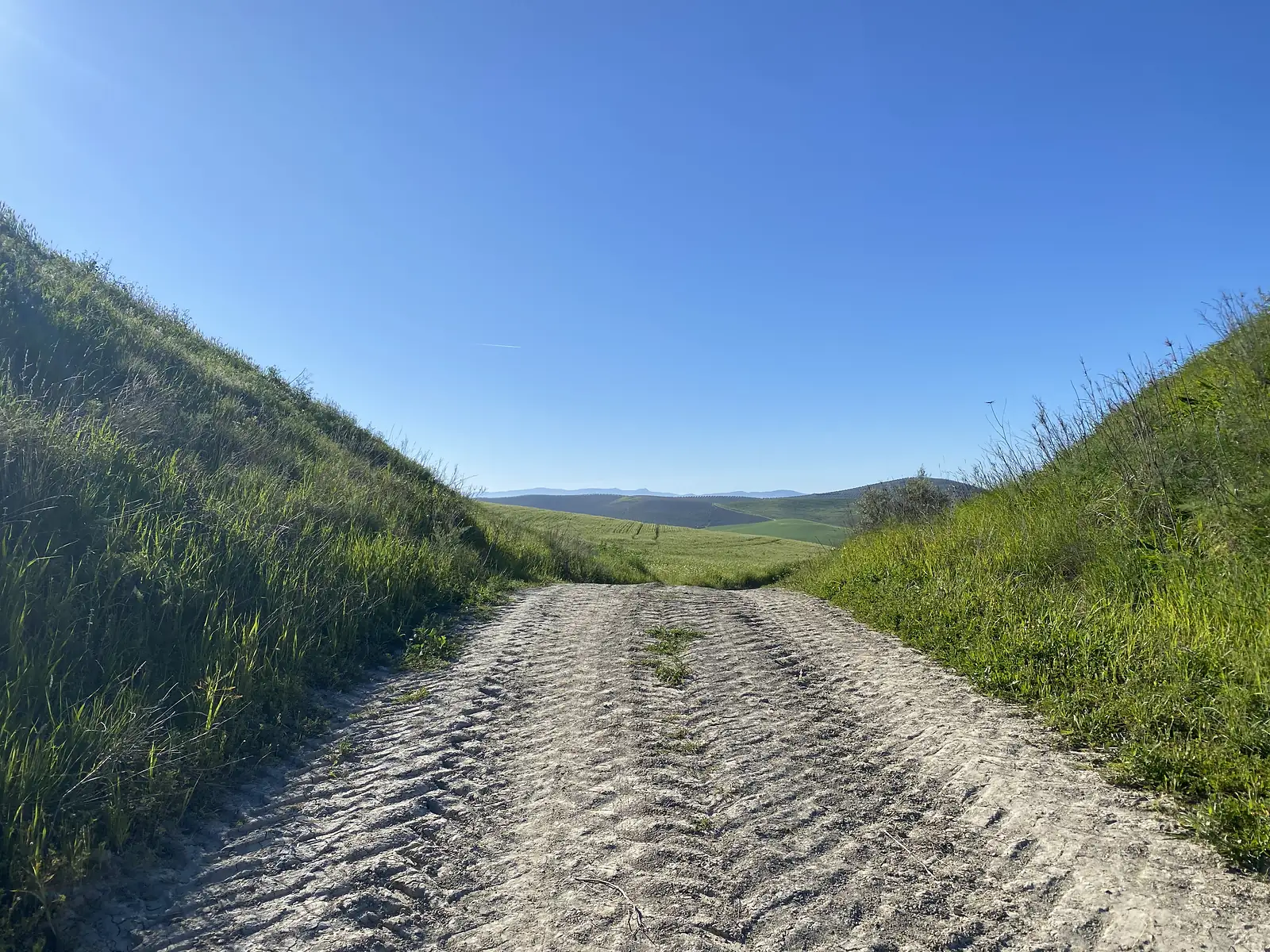 A sandy double track dipping between steep grassy banks towards rolling green fields and hazy blue hills.