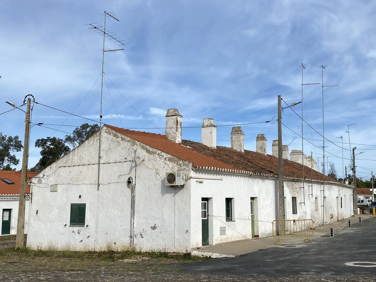A long whitewashed single-storey terrace with a red pantile roof and a row of tall chimneys, TV aerials above it and yellow barriers on the pavement.