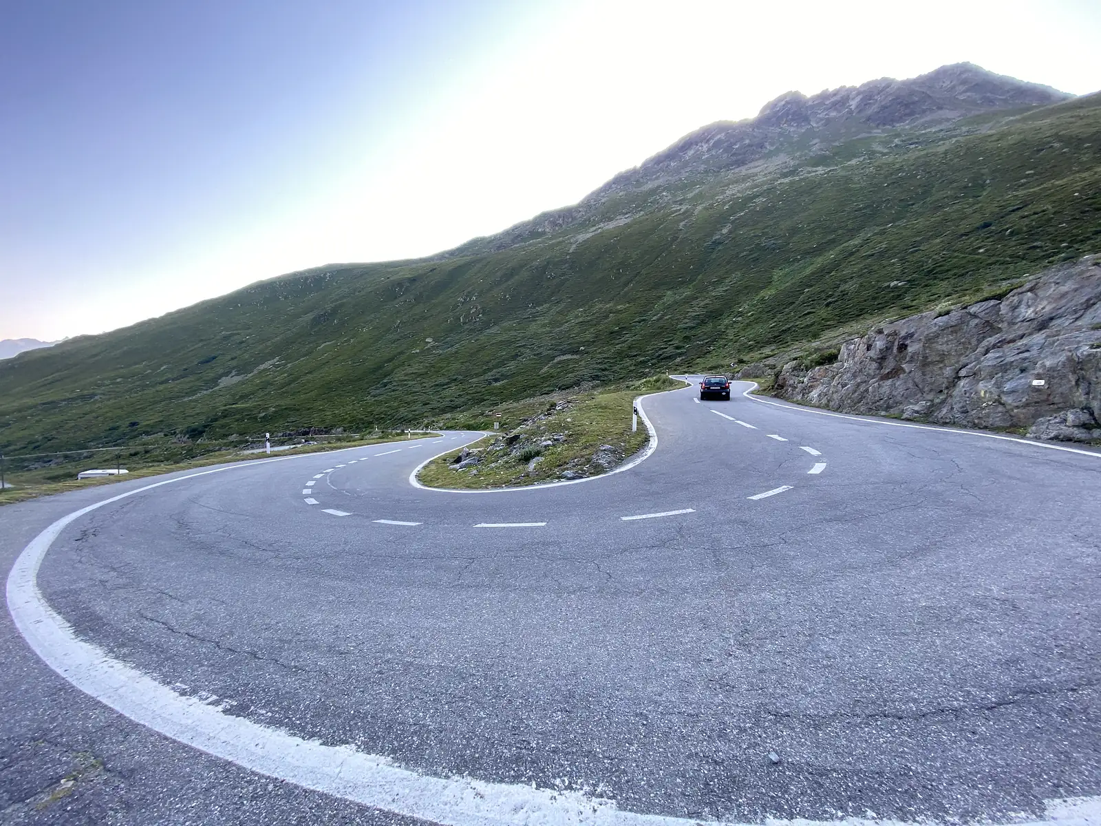 A car rounds a sharp hairpin bend on a mountain road, with grassy slopes and a rocky outcrop rising above.
