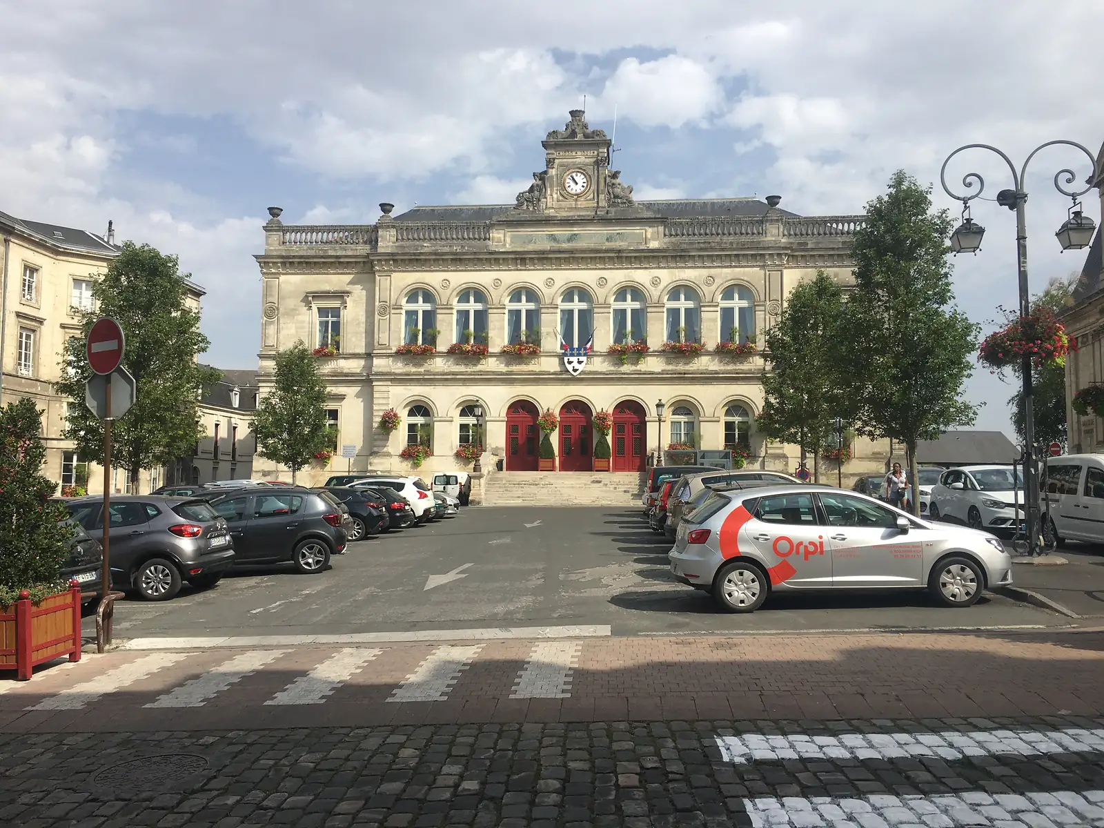 An ornate stone town hall with a central clock tower overlooks a paved square lined with parked cars and trees.