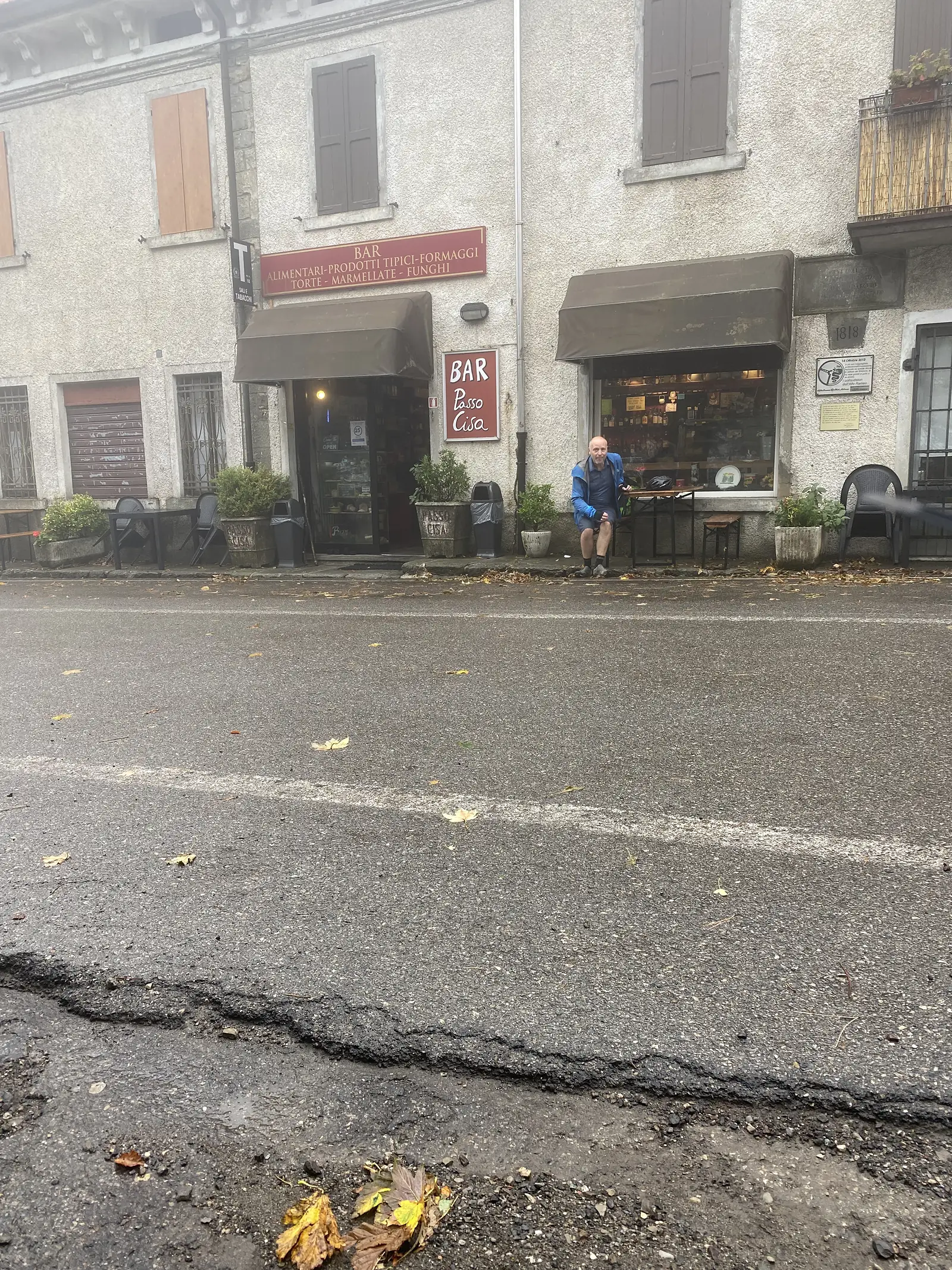 A man in cycling kit stands outside a shopfront signed BAR Passo Cisa, alongside a red sign advertising Alimentari, prodotti tipici, formaggi, torte, marmellate, funghi.