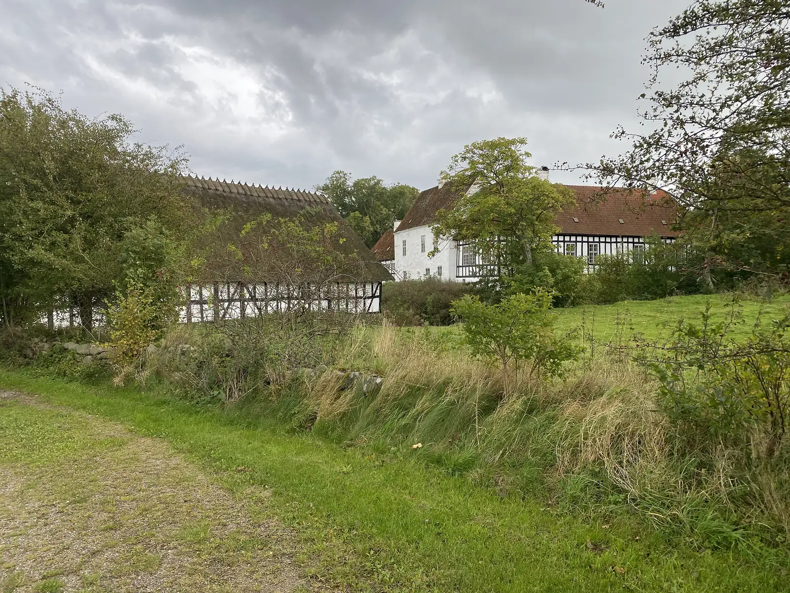 A gravel track past a hedge bank to a thatched half-timbered farm range and a white manor with a red-tiled roof under heavy cloud.
