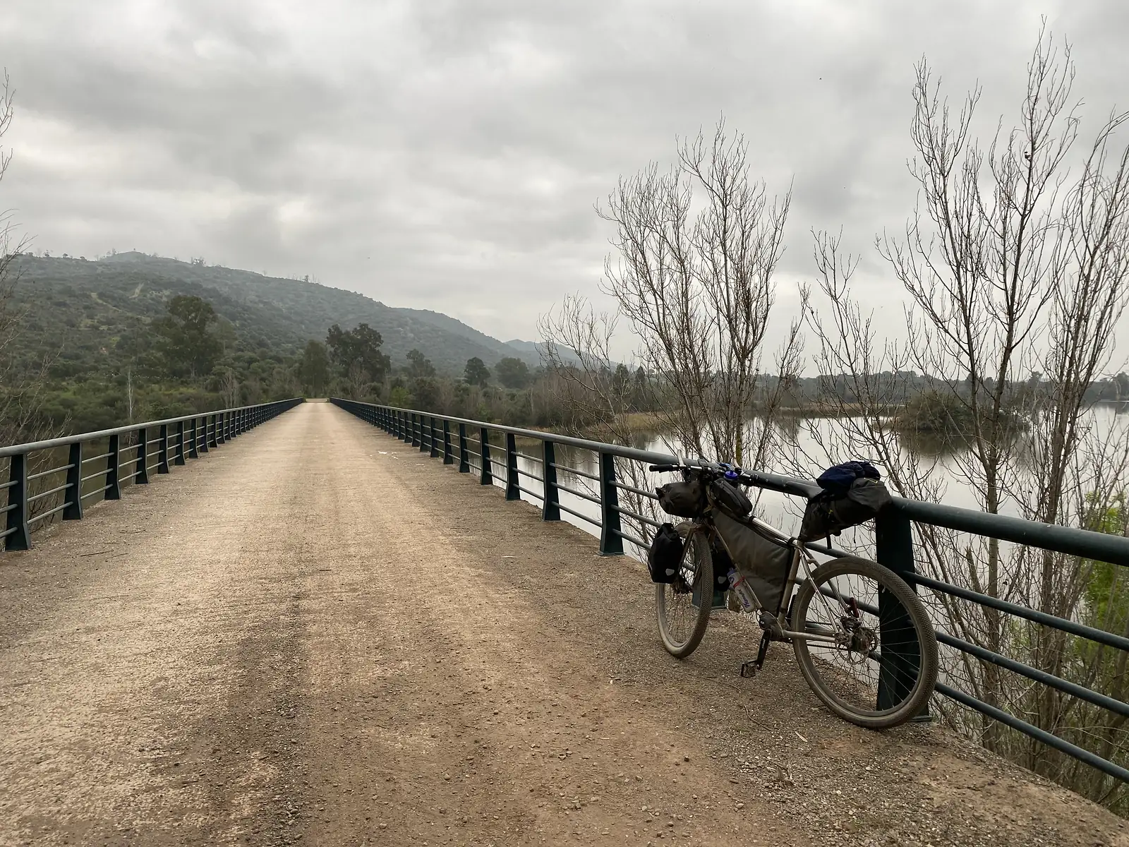 A loaded bikepacking bike leaning against the green railing of a long straight bridge carrying a gravel path across still brown water.