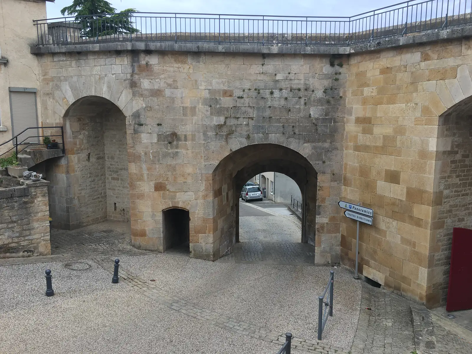 A stone archway through the Langres town walls opening onto a cobbled street, with a directional sign for Panoramics and Toutes Directions beside it.