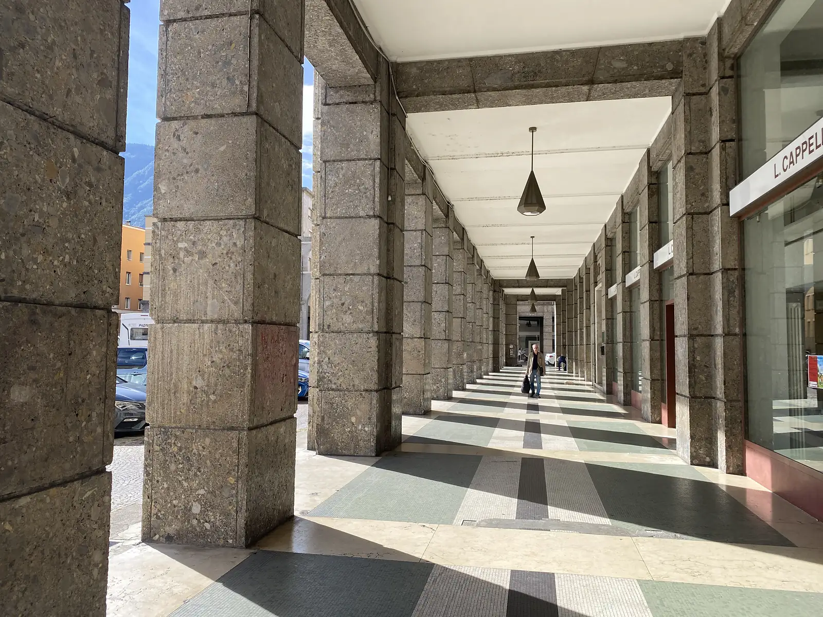 A long arcade of heavy stone piers runs beside shopfronts over a chequered marble floor, a single figure walking towards the far end.