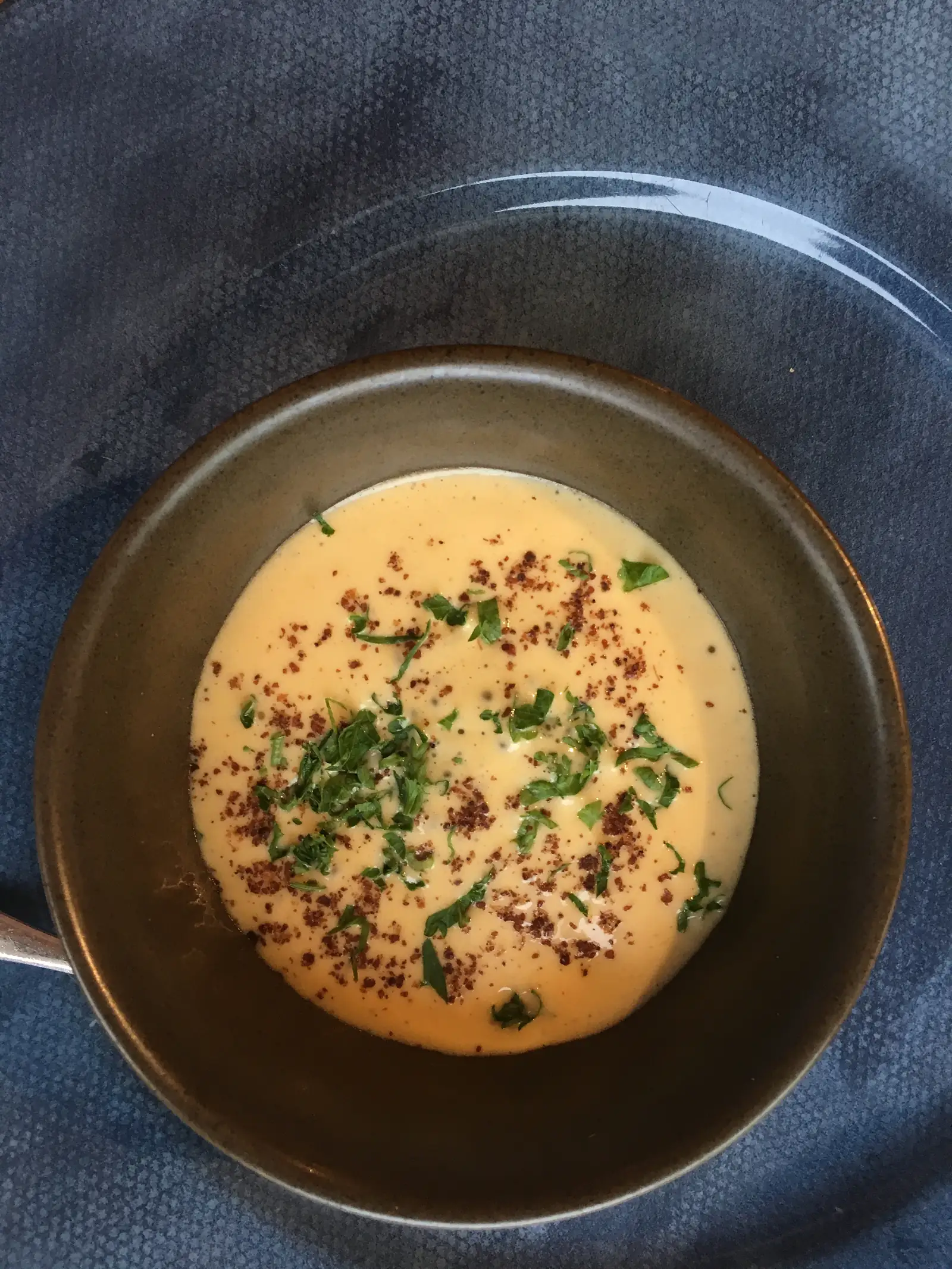 A pale, creamy soup in a dark bowl, topped with chopped herbs and crisp savoury crumbs.