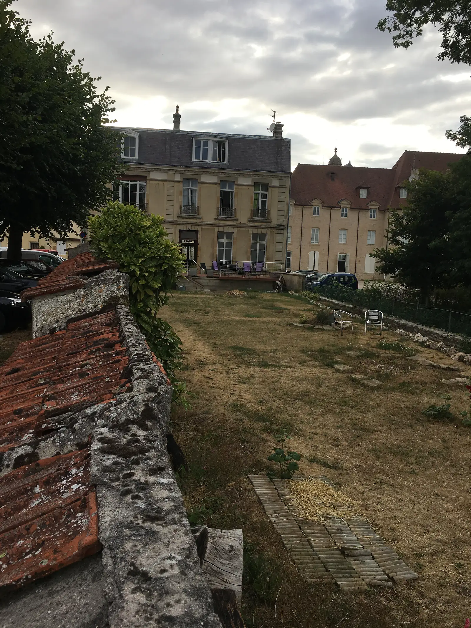 A church spire rising behind pale stone townhouses in Langres, seen across an overgrown garden from beside a weathered tiled wall coping.