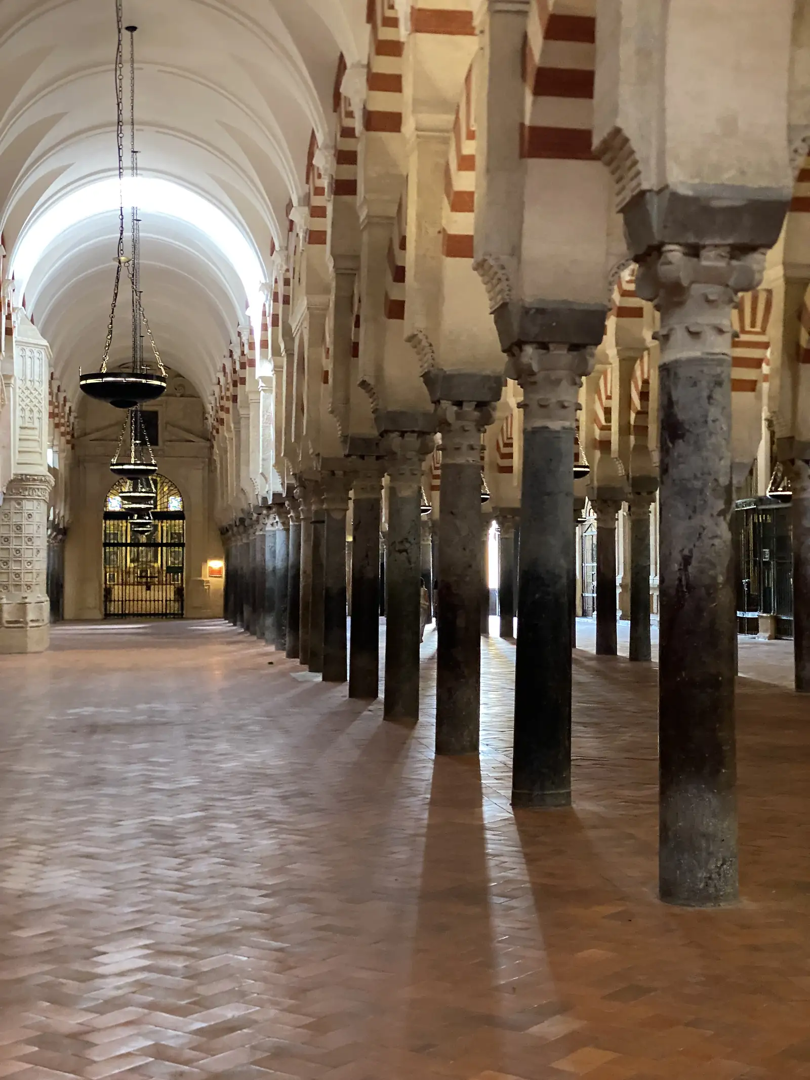 A long aisle of dark marble columns and striped arches with hanging bowl lamps above a herringbone brick floor.