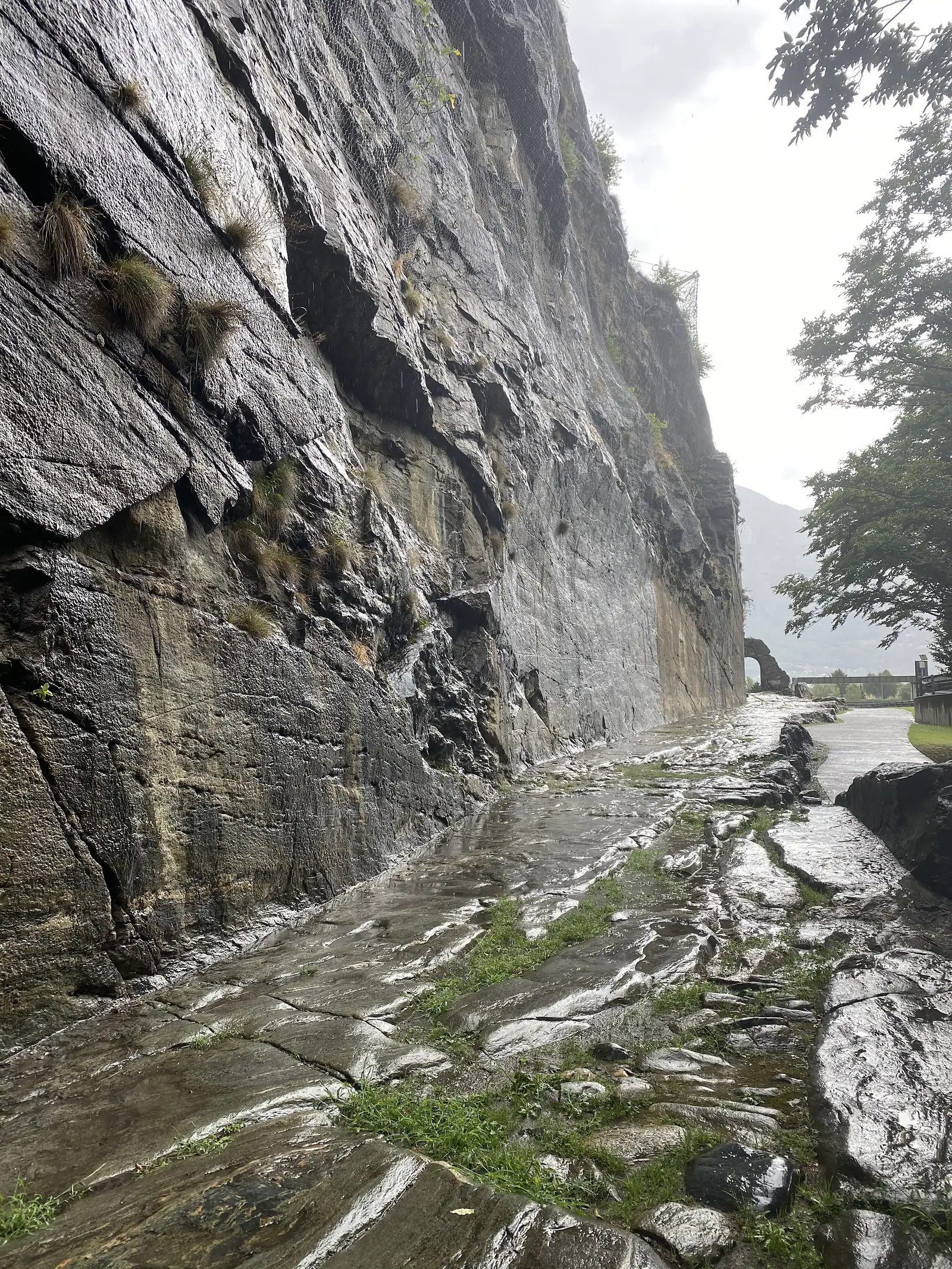 A rain-streaked path running below a sheer cut rock face, with a stone archway and trees visible further along the route.