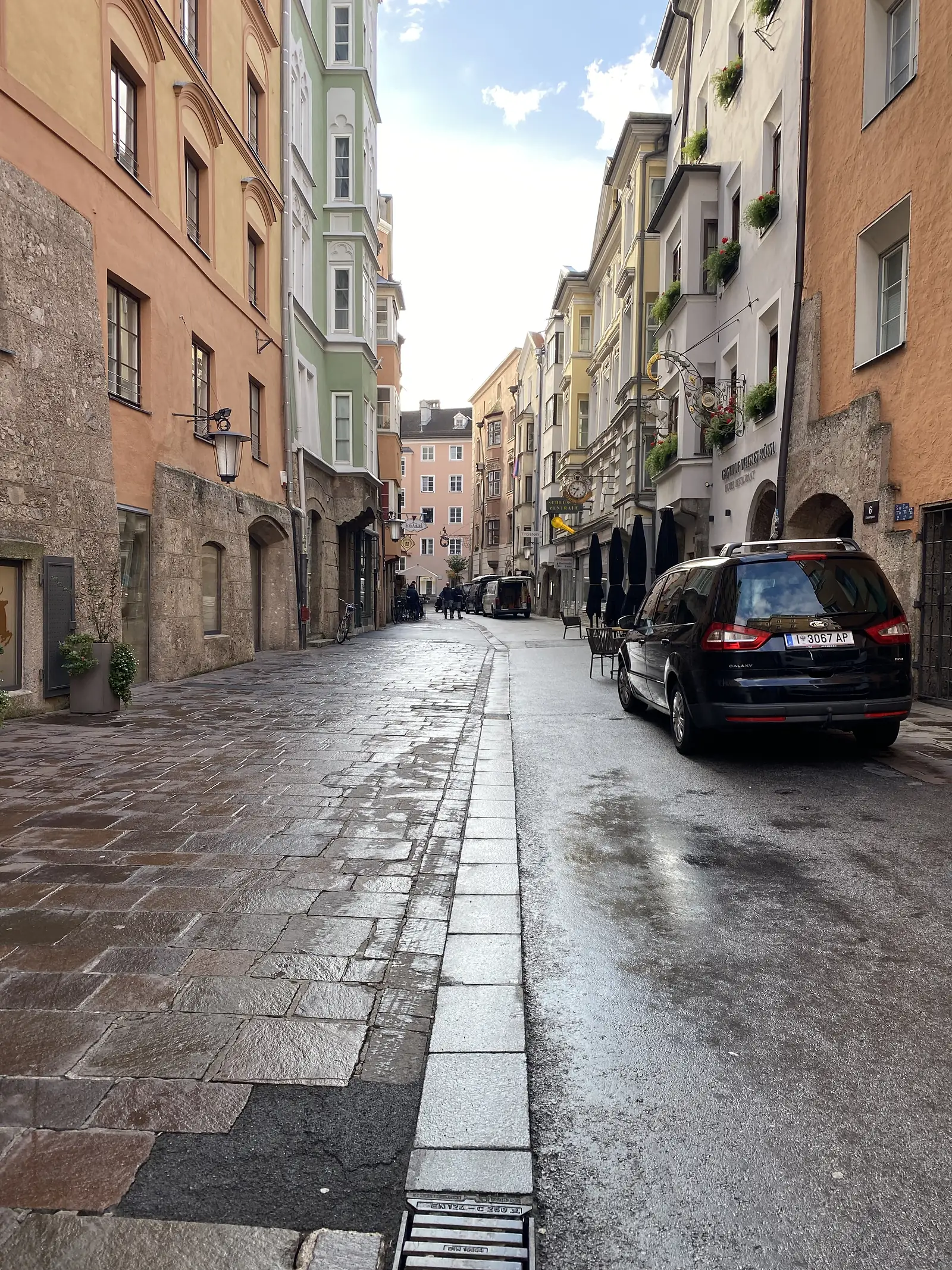 A wet cobbled street winds between pastel-painted old-town buildings with café tables and umbrellas set out at the kerb.