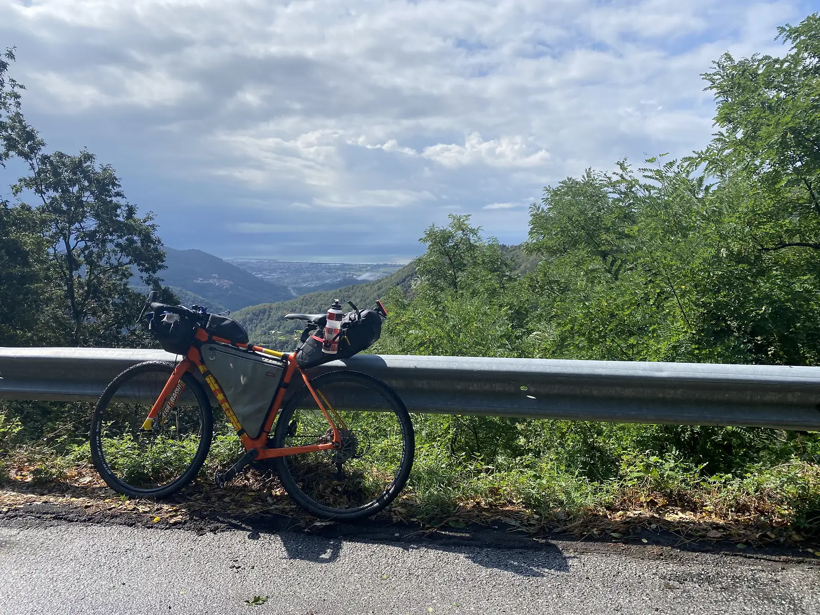 A loaded orange gravel bike leaning against a roadside guardrail, with wooded hills falling away to a hazy coastal town and the sea beyond.