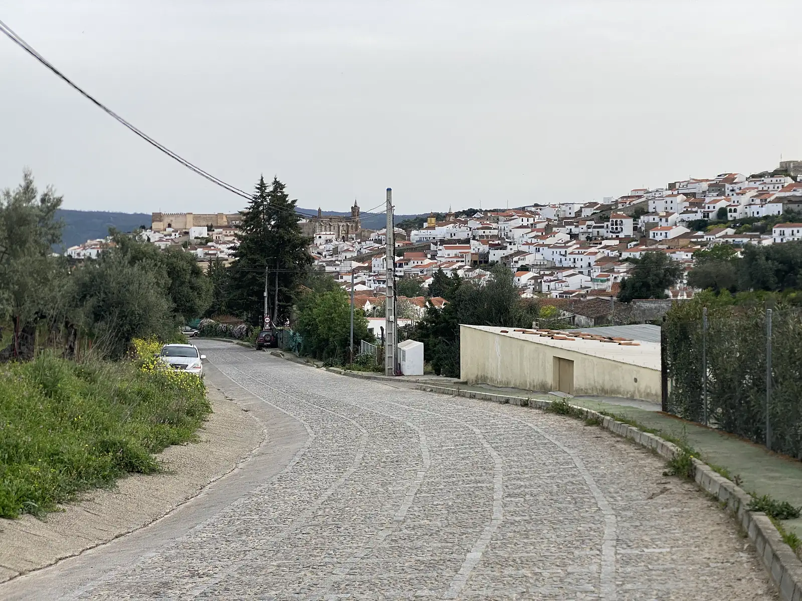 A cobbled road curving downhill towards a hillside town of white houses and red roofs, with a castle and a church tower along its skyline.