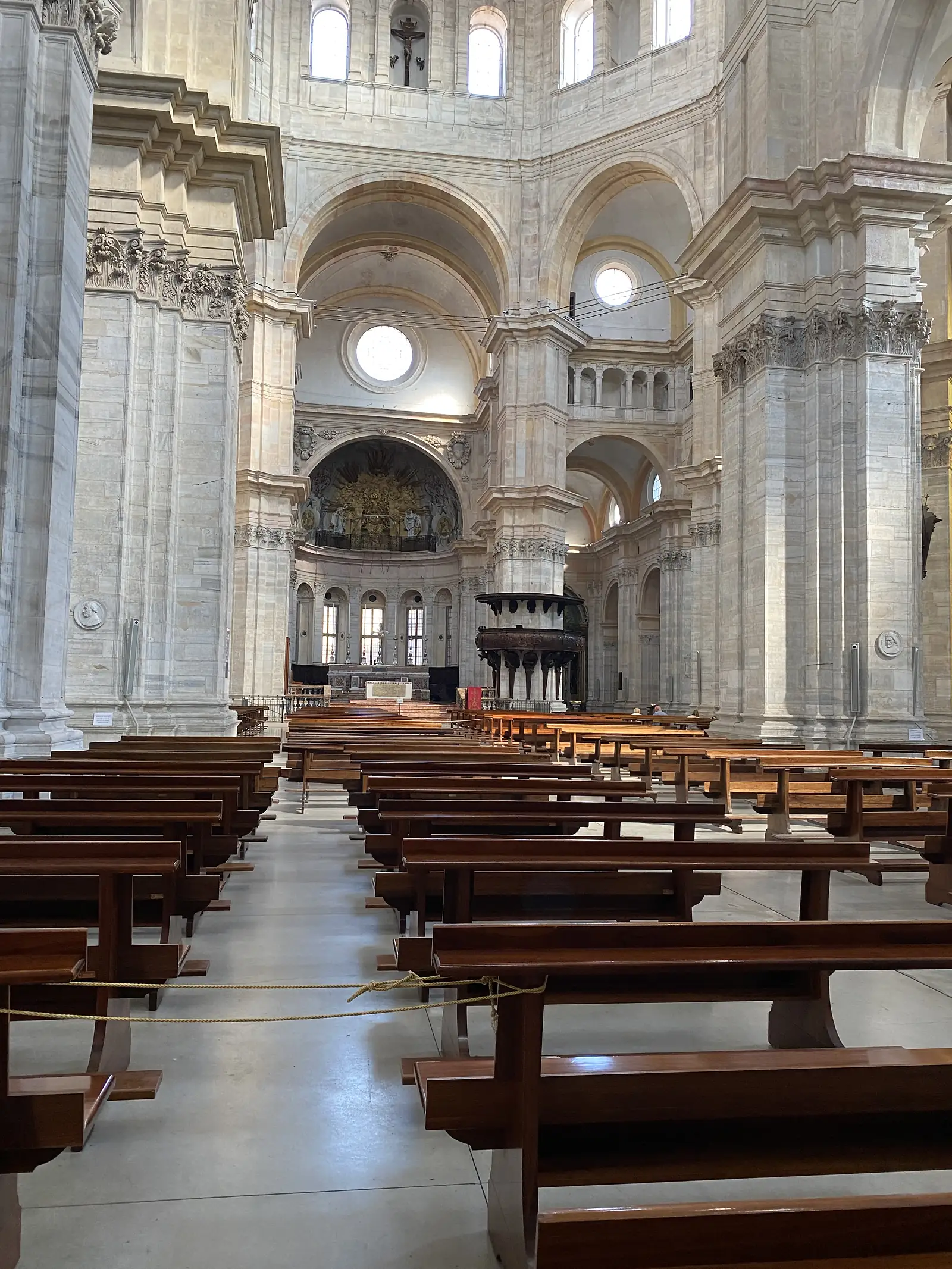 Rows of wooden pews fill the marble nave of a grand domed church interior, its side chapels lit by round windows and gilded frescoes.