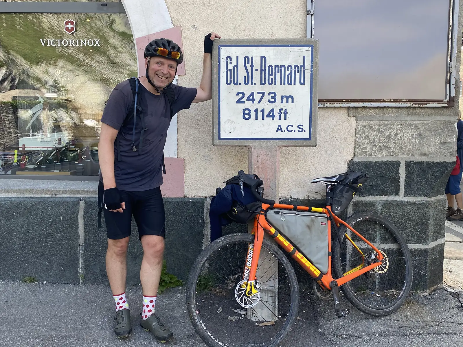 The same cyclist smiles beside his orange bikepacking bike at the 'Gd. St-Bernard 2473m' summit marker, with other visitors passing nearby.