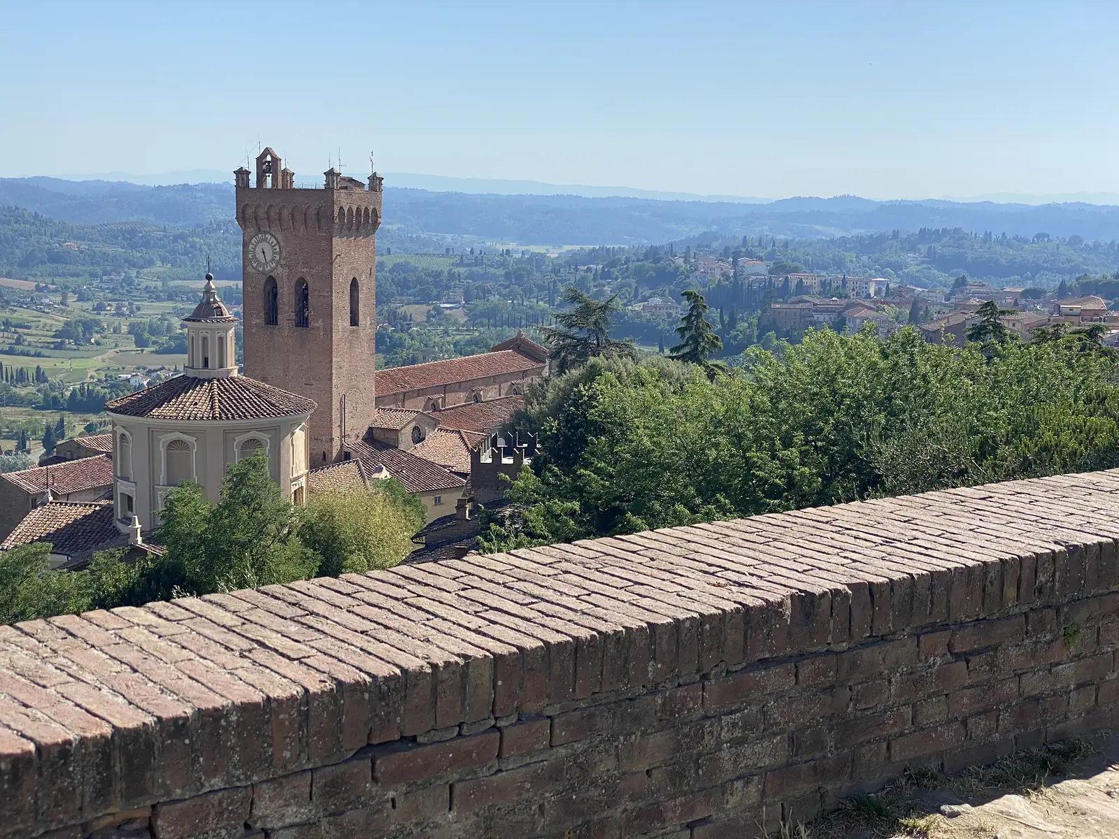 A brick-crenellated town wall looks over a domed church and a tall crenellated clock tower with bells, with rolling wooded hills stretching into the distance beyond the rooftops.