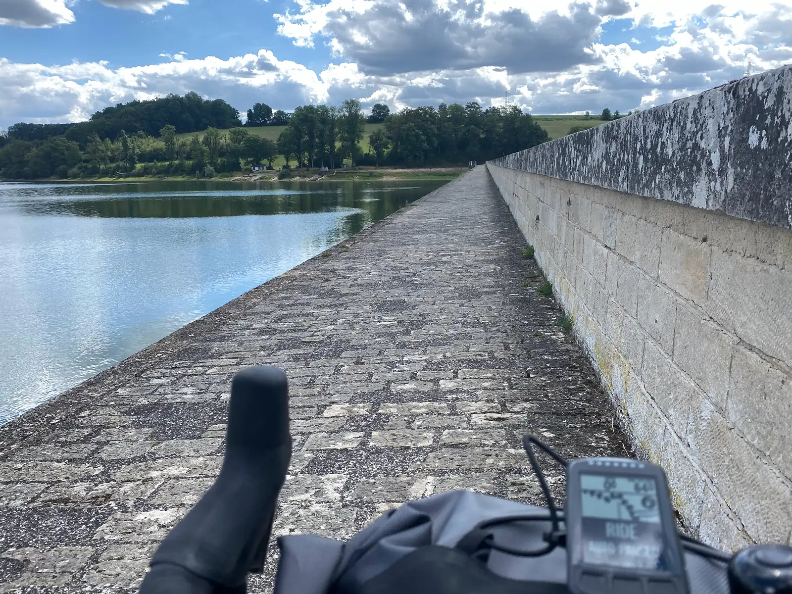 A cyclist's view along the cobbled top of a long stone dam beside a reservoir, handlebar bag and GPS computer visible in the foreground.