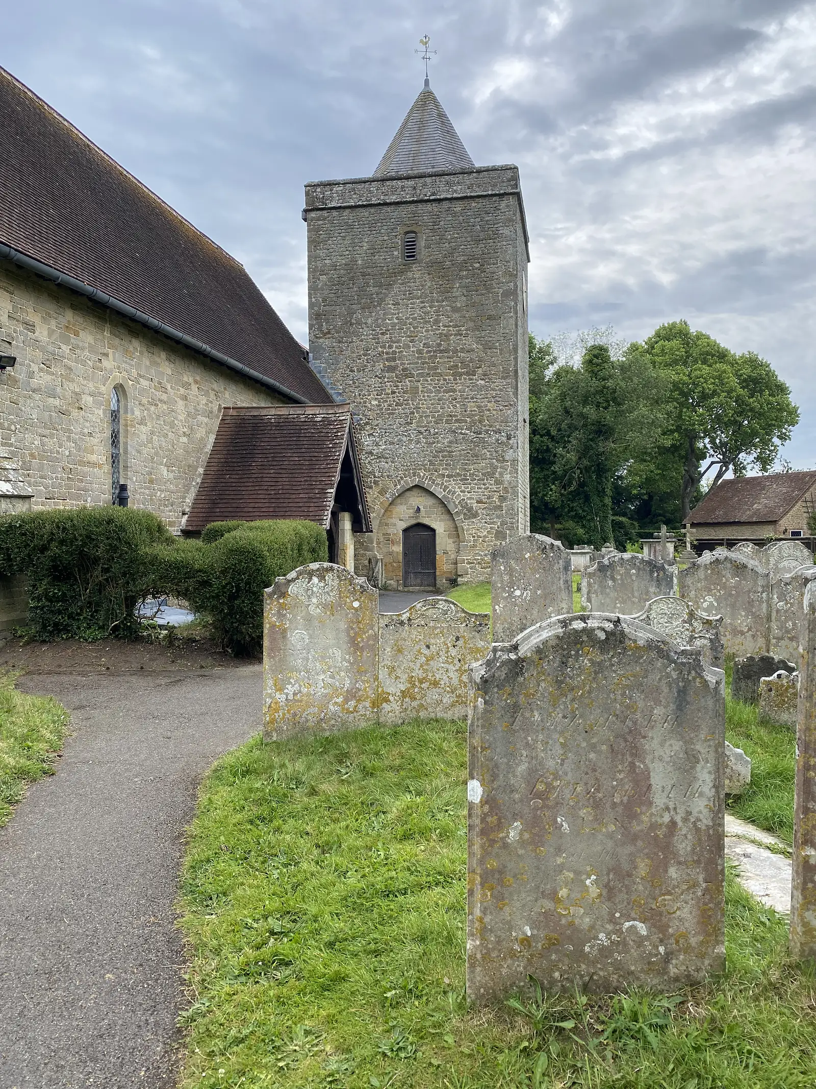 A stone church with a shingled spire on a squat tower and a tiled porch, seen across lichened headstones.