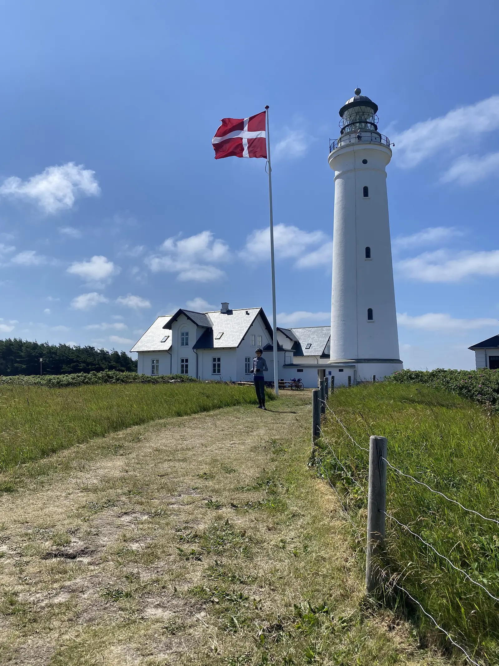 A white lighthouse beside a gabled keeper's house, the red-and-white Danish flag flying from a mast and a person standing on the grass path below.