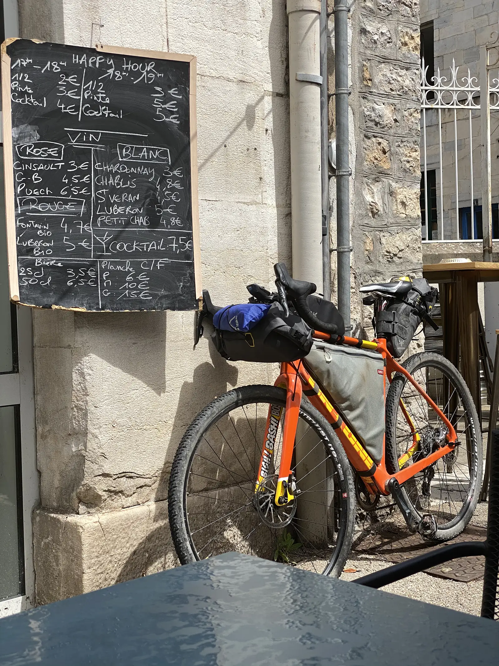 A loaded orange bikepacking bike rests against a stone wall beside a handwritten blackboard listing happy-hour drink prices outside a bar.