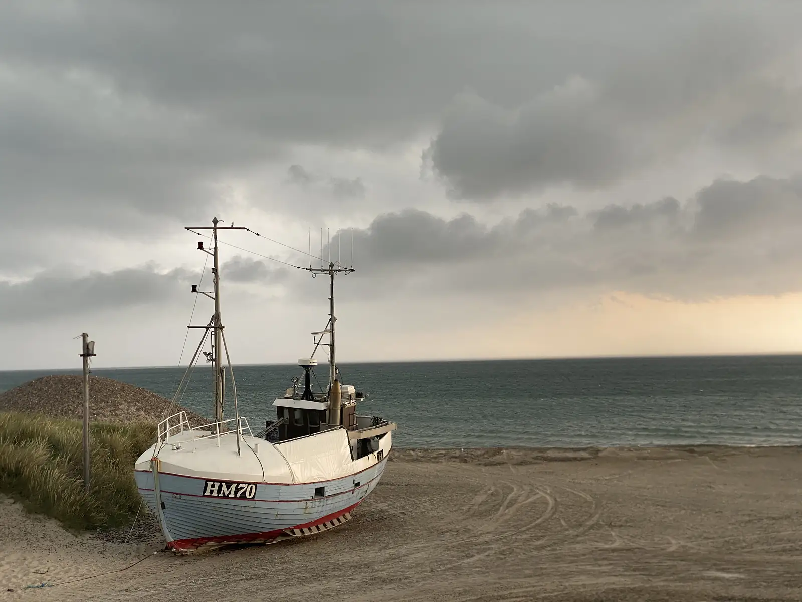 A single-masted fishing boat lettered "HM70" hauled up on the sand under heavy cloud, with a band of pale sunset over the sea.