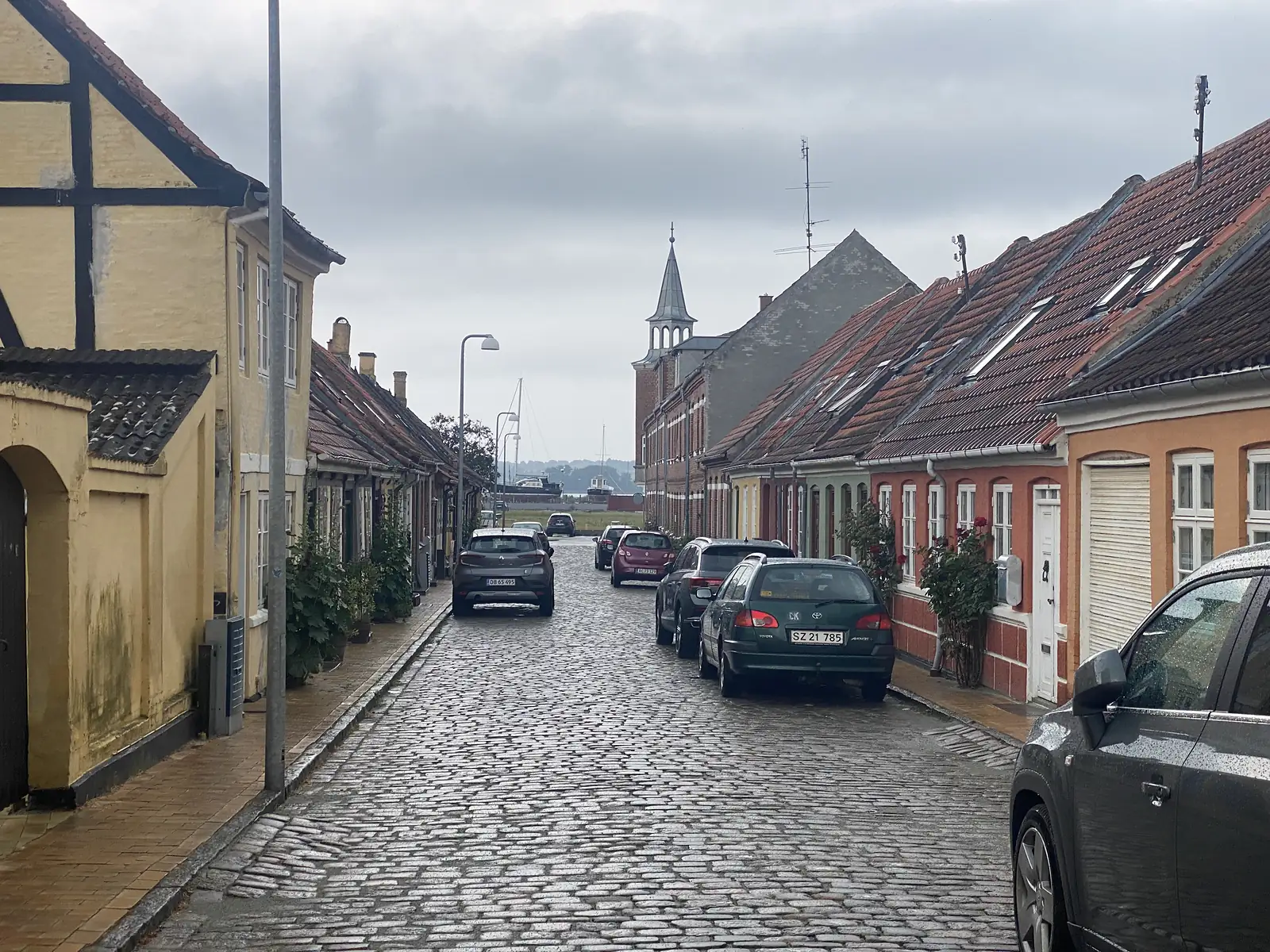 A wet cobbled street of yellow half-timbered and red-brick houses with hollyhocks, sloping down towards a harbour of masts and a red brick spire.