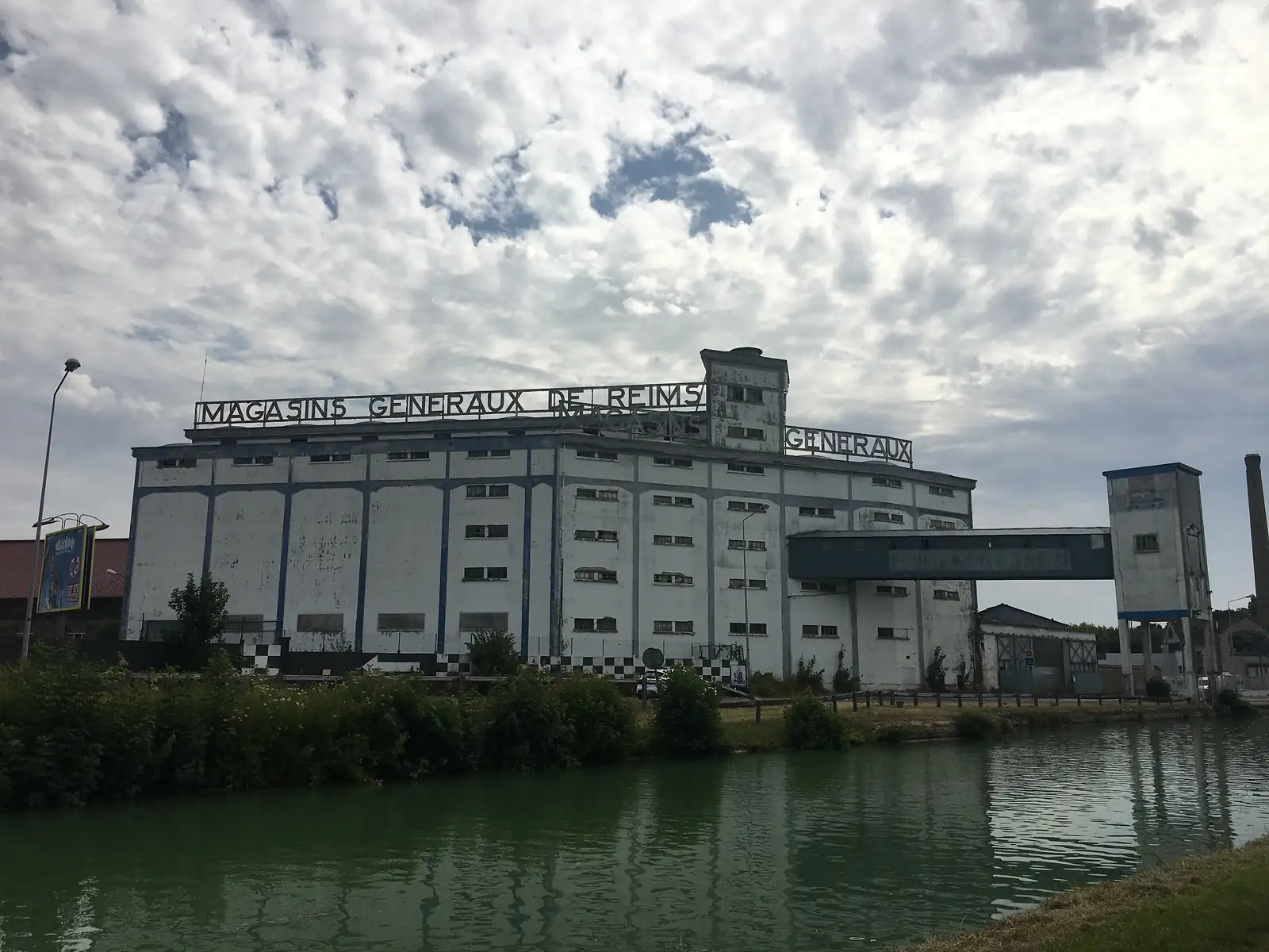 The disused "Magasins Généraux de Reims" grain warehouse beside a canal, its long white facade lettered with the building's name below a rooftop water tower.