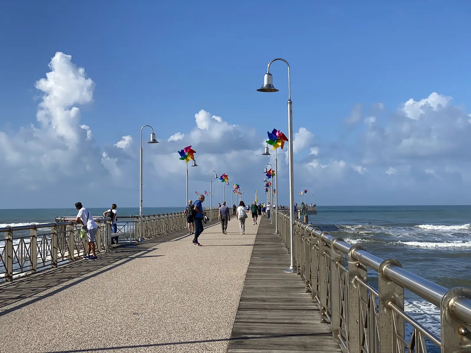 Pedestrians walk along a pier lined with lamp posts decorated with colourful pinwheels, the sea stretching away on both sides under a bright blue sky.