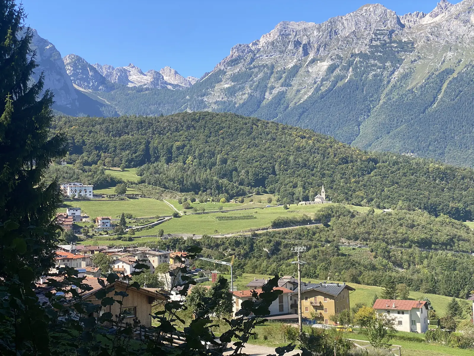 A village of pale houses and a slim church spire sits on a green shelf below jagged grey peaks, a construction crane rising among the rooftops.