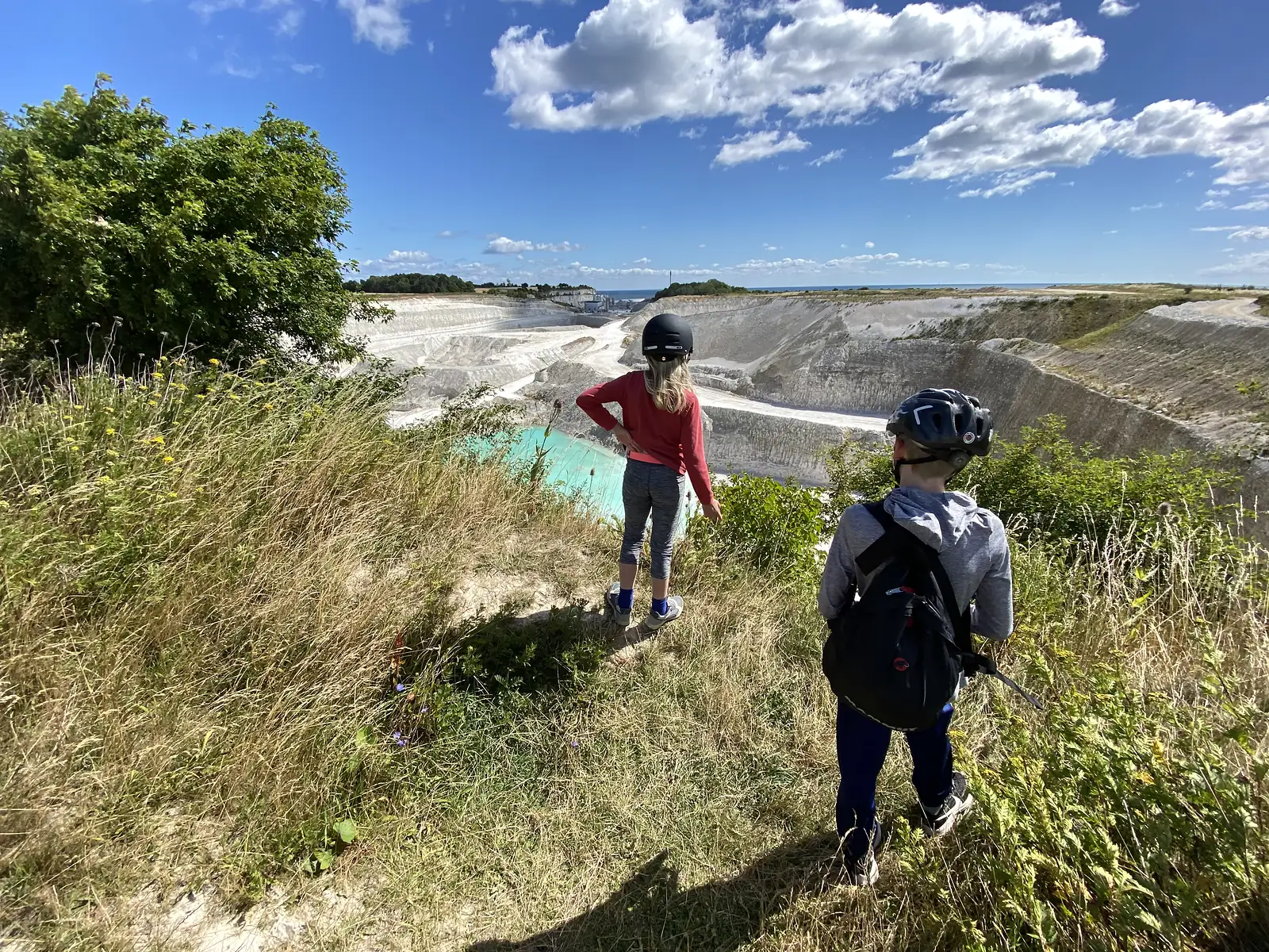 Two children in cycling helmets on a grassy rise looking down into a huge white chalk quarry with a turquoise pool, the sea beyond.