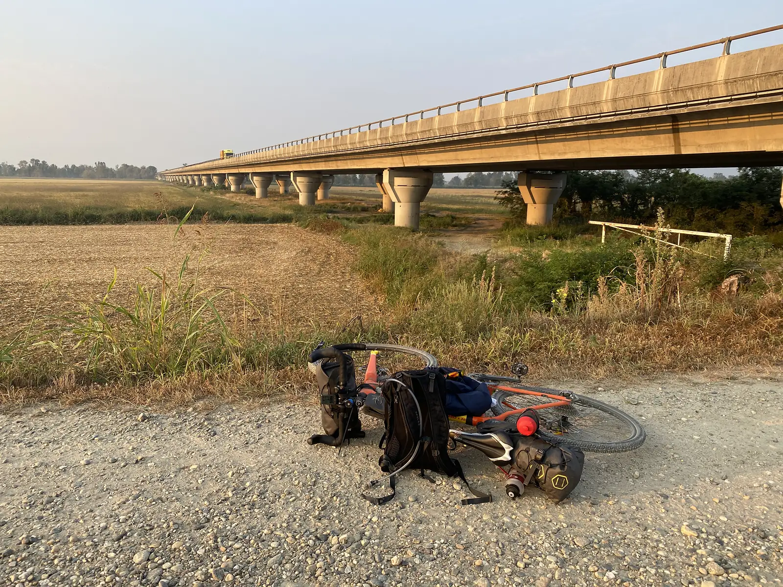A loaded touring bike lies stripped of its front wheel on a gravel verge beneath a long concrete viaduct crossing open farmland.