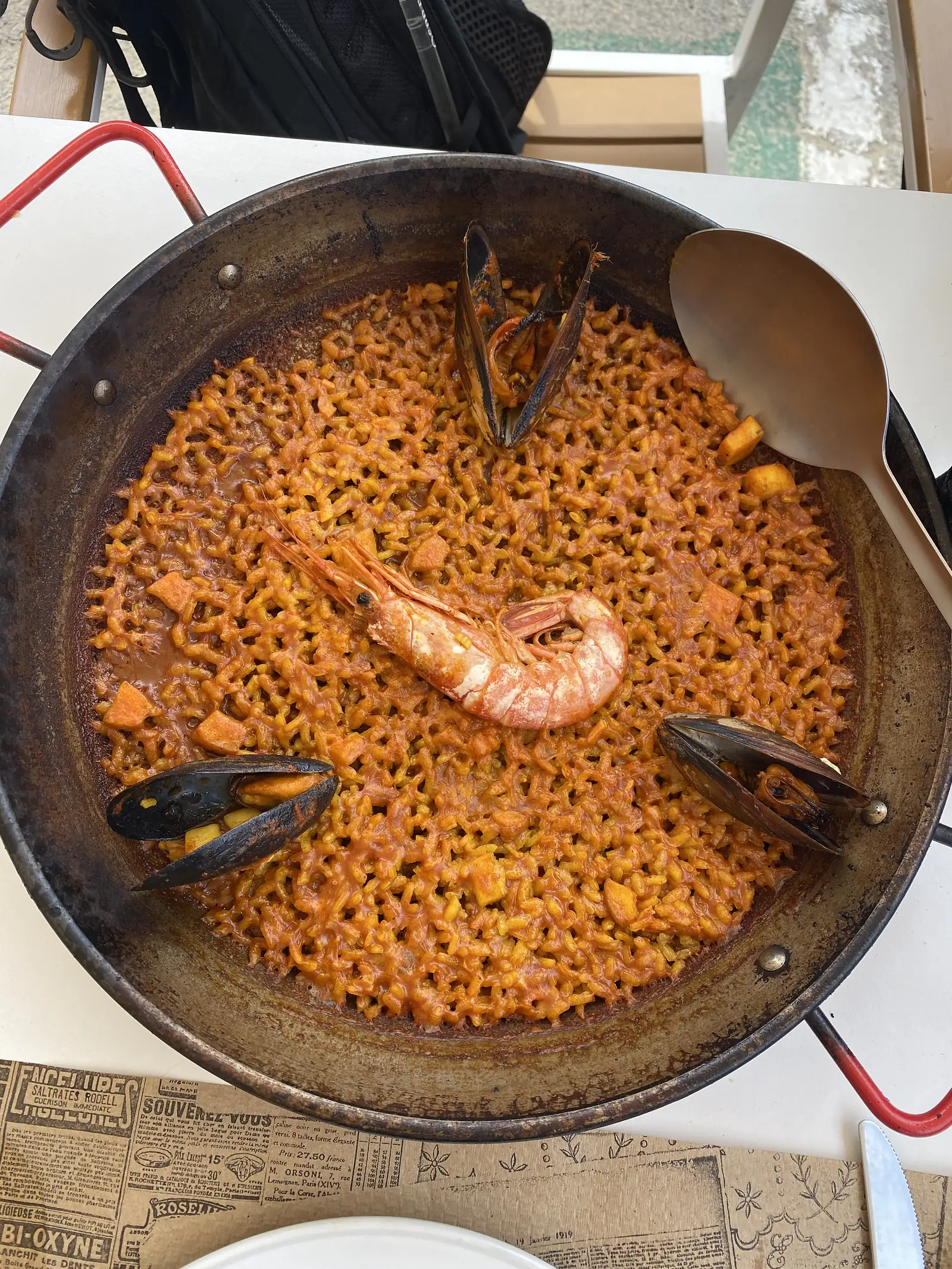 A shallow steel pan of orange-red short-noodle fideua topped with a whole prawn and mussels, with a serving spoon.