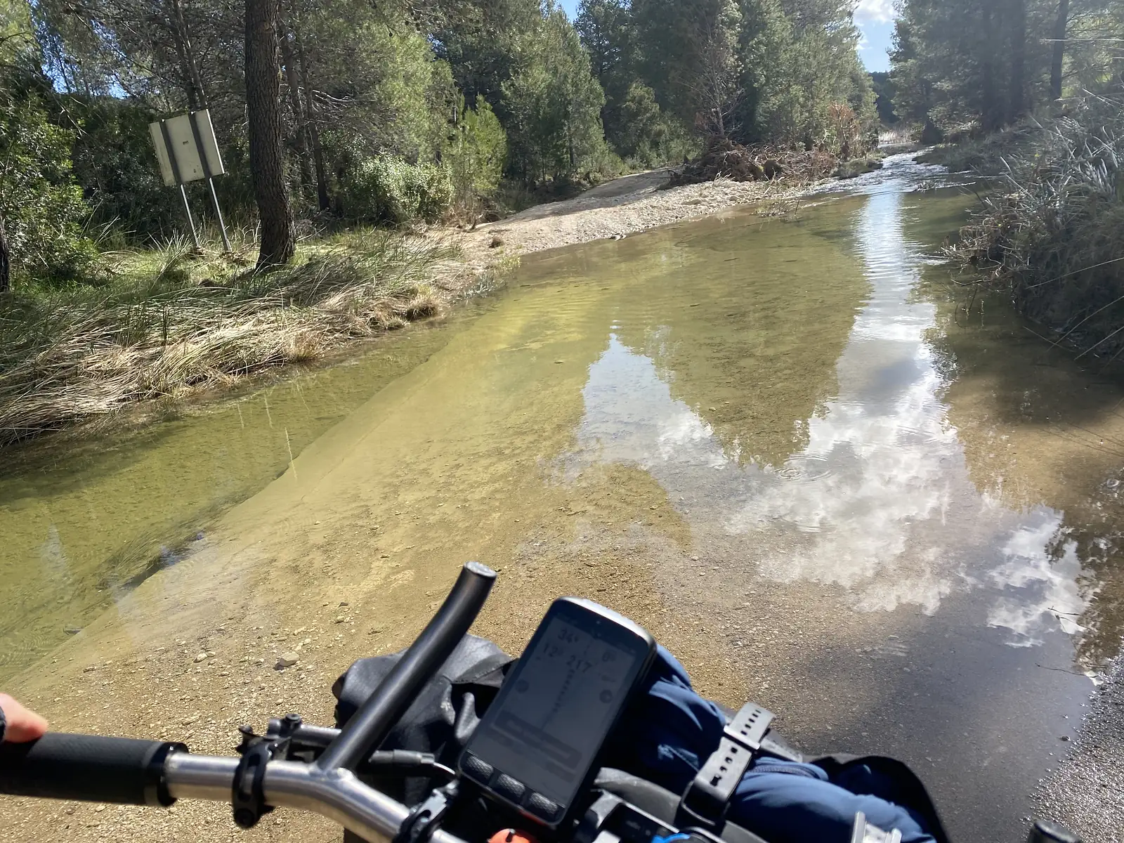 A flooded gravel track seen over the handlebars, a shallow stream running down the road through pine woods.