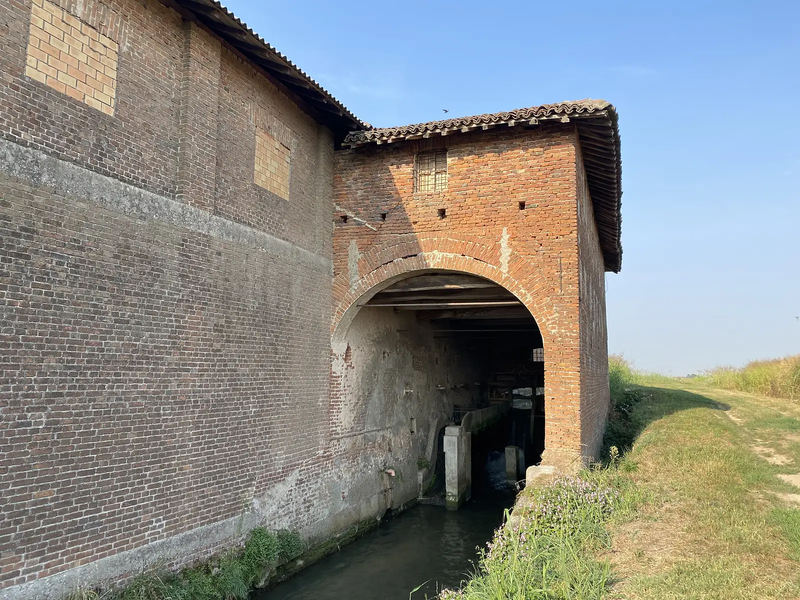 A weathered brick farmhouse with a tiled roof straddles a stream, its arched opening framing the water flowing beneath.