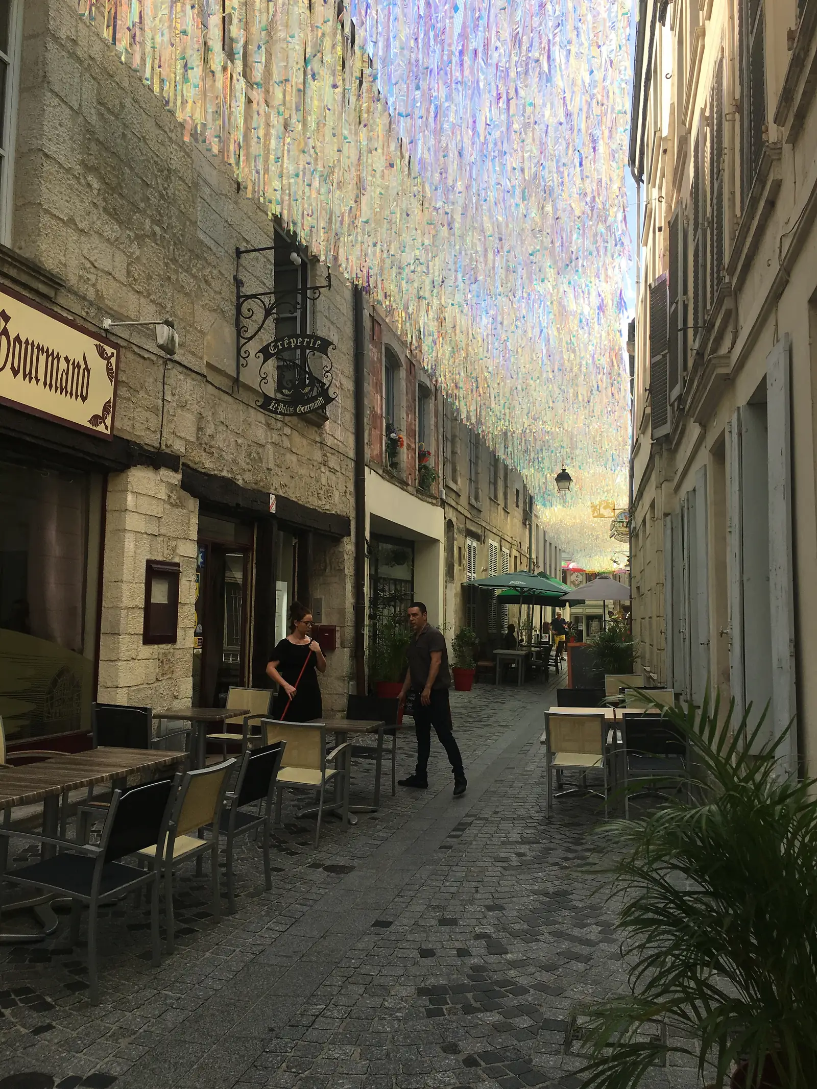 A narrow cobbled street lined with café tables and hanging flower baskets leads past a sign for a 'Crêperie', its stone walls glowing in the afternoon sun.