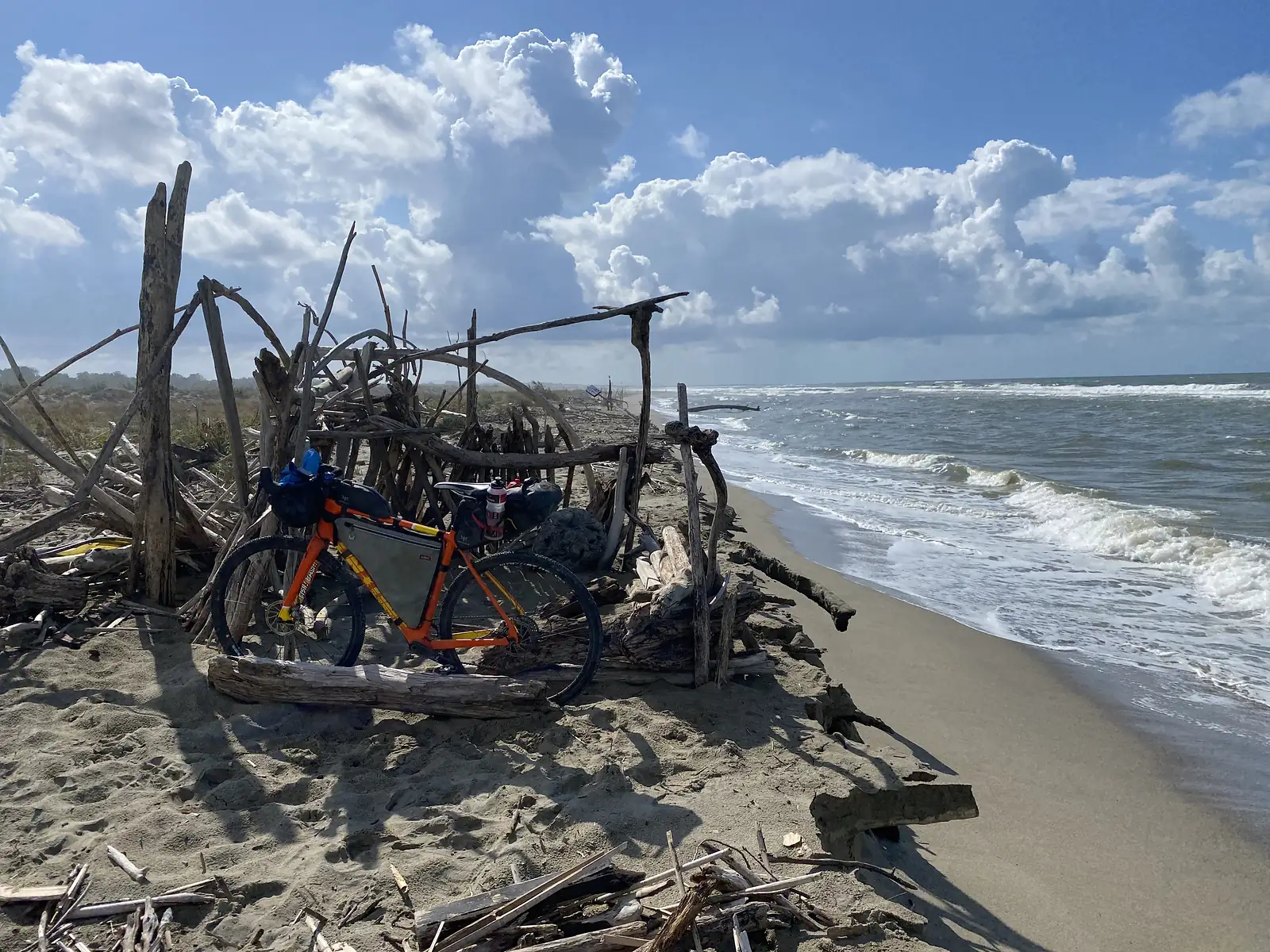 The loaded orange bike leaning inside the driftwood shelter, with a distant beach sign and the sea beyond.
