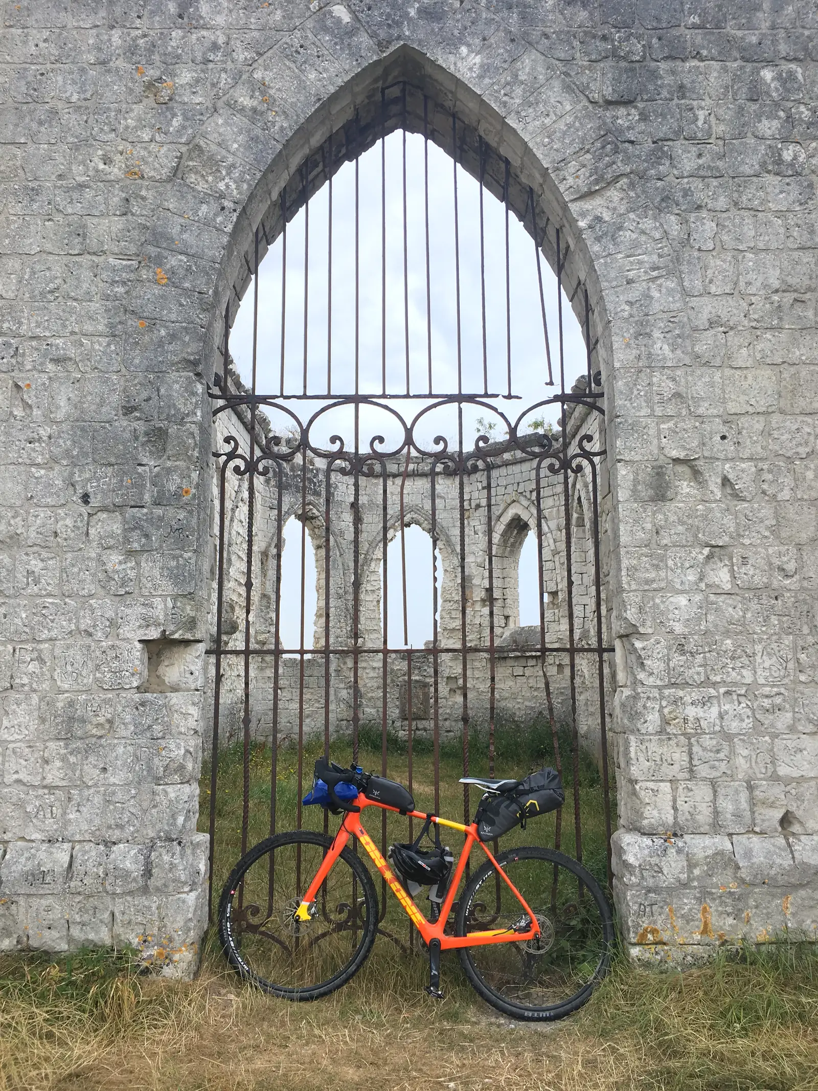 Looking through the wrought-iron gate of a ruined chapel's arched doorway towards its bare stone interior, with a loaded orange gravel bike parked outside on the grass.