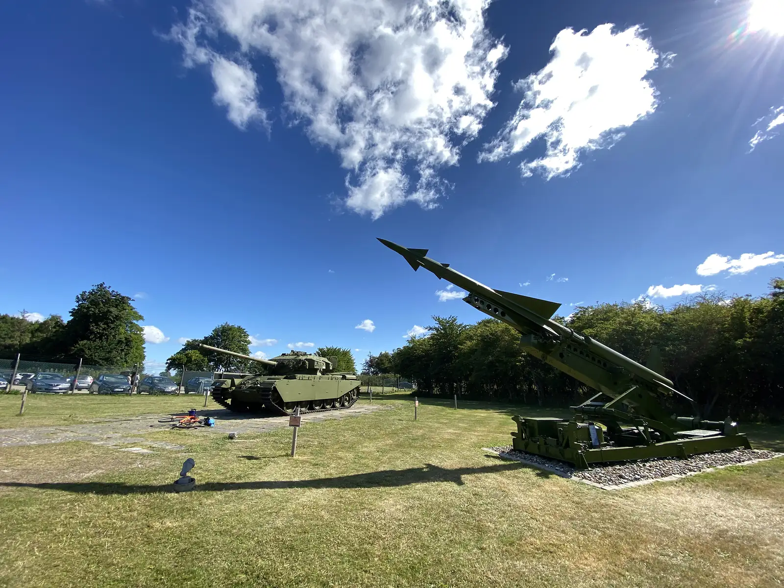 A tank and a missile on its launcher displayed on grass at an outdoor military museum, with bikes laid on the ground nearby.