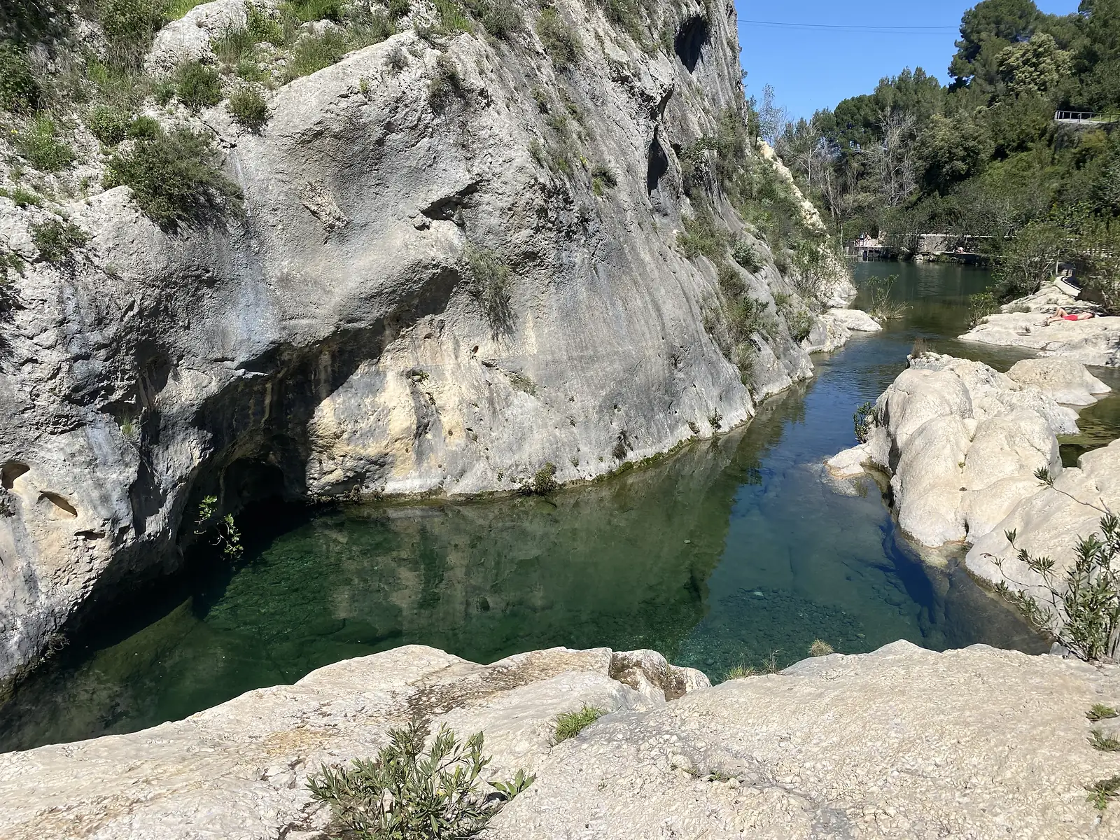 A deep green river pool held between smooth limestone cliffs and rock slabs, with a footbridge and people at the far end.