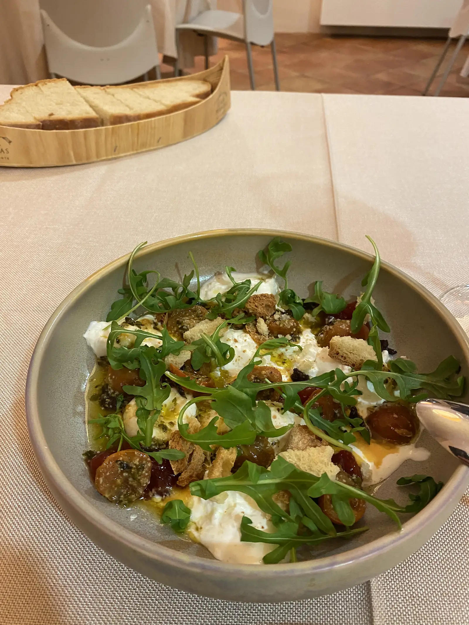 A bowl of burrata with cherry tomatoes, rocket and croutons in green oil, a wooden tray of sliced bread on the table behind.