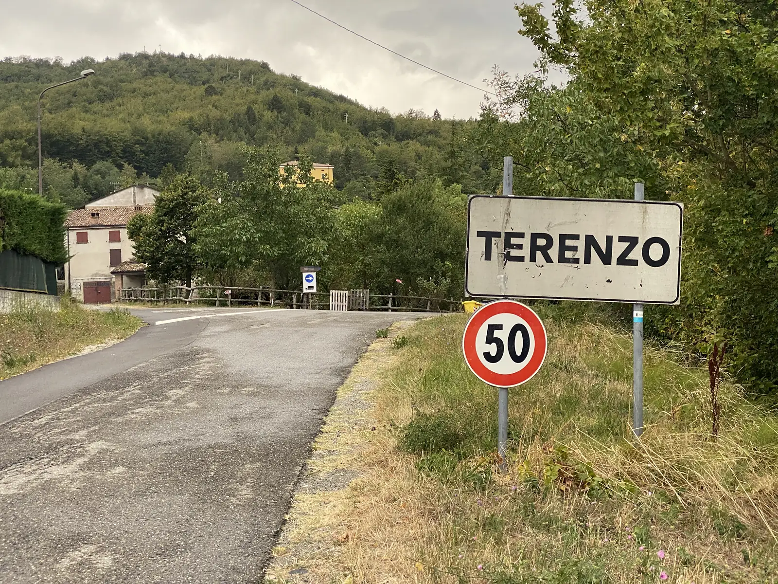 A village-entry sign reading 'Terenzo' with a 50 km/h speed-limit disc stands beside a quiet lane running past a stone farmhouse and wooden gate.