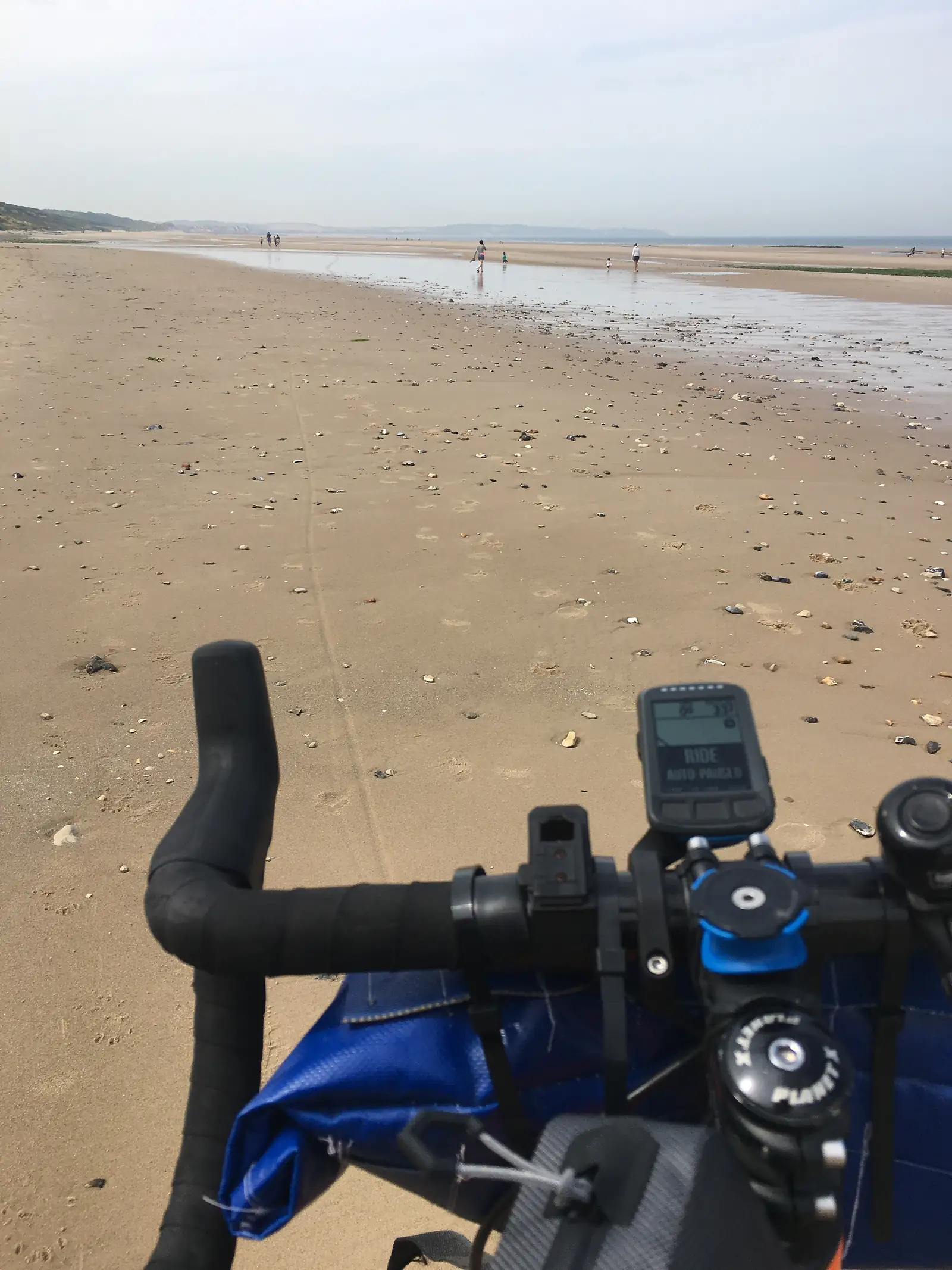A bike computer mounted on the handlebars reading 'RIDE AUTO PAUSED', looking out over a pebble-strewn beach with walkers in the distance.