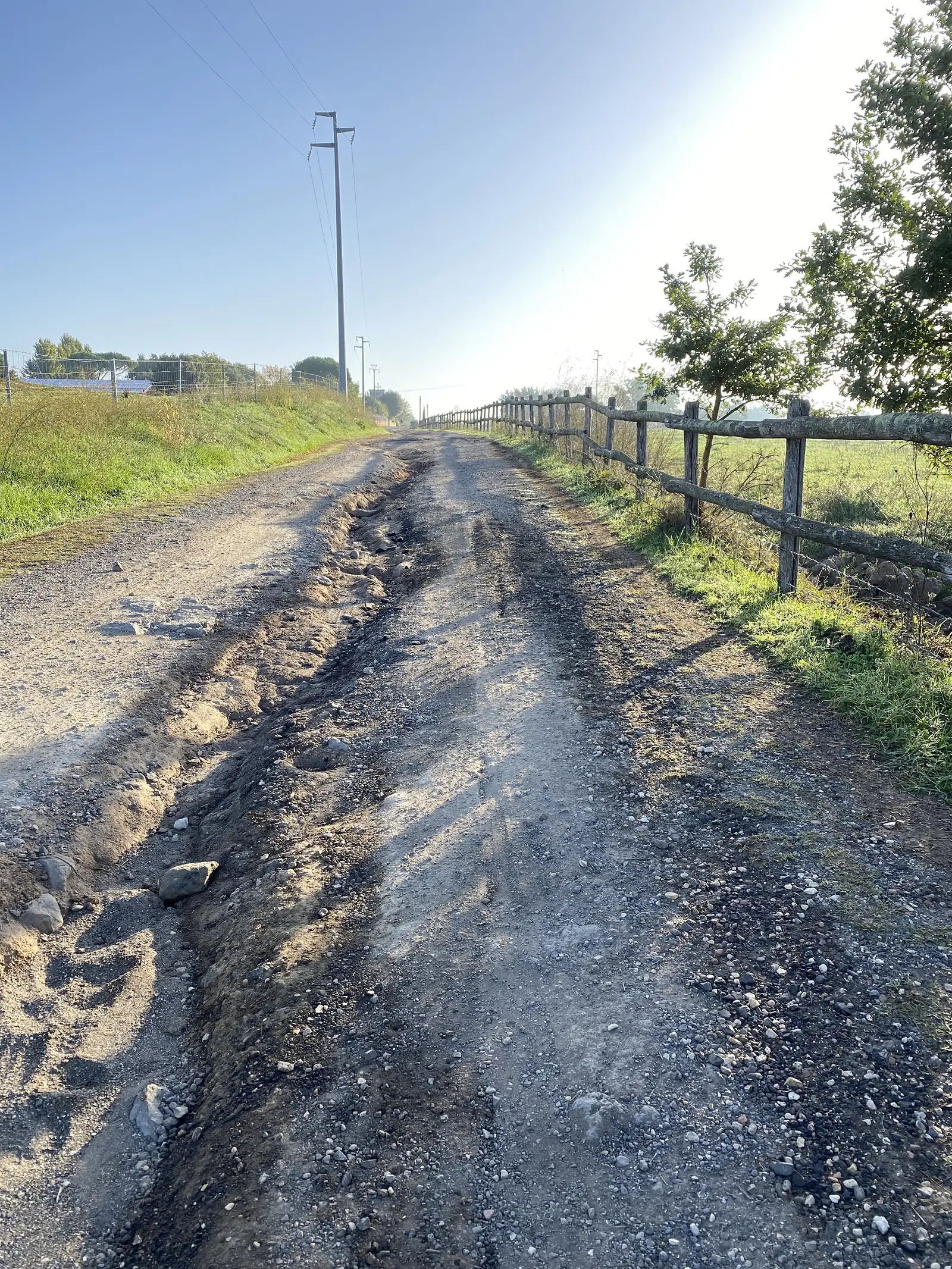 A pale stony track stretching into the distance beside a wooden post fence, with an electricity pylon rising over the fields ahead.