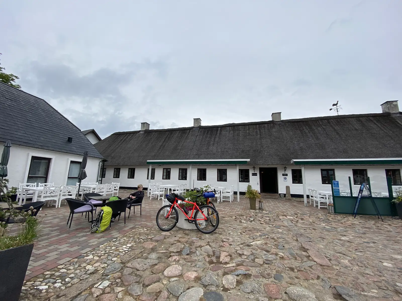 A loaded red bike propped on a cobbled courtyard before a long white thatched inn with green awnings and white cafe furniture.