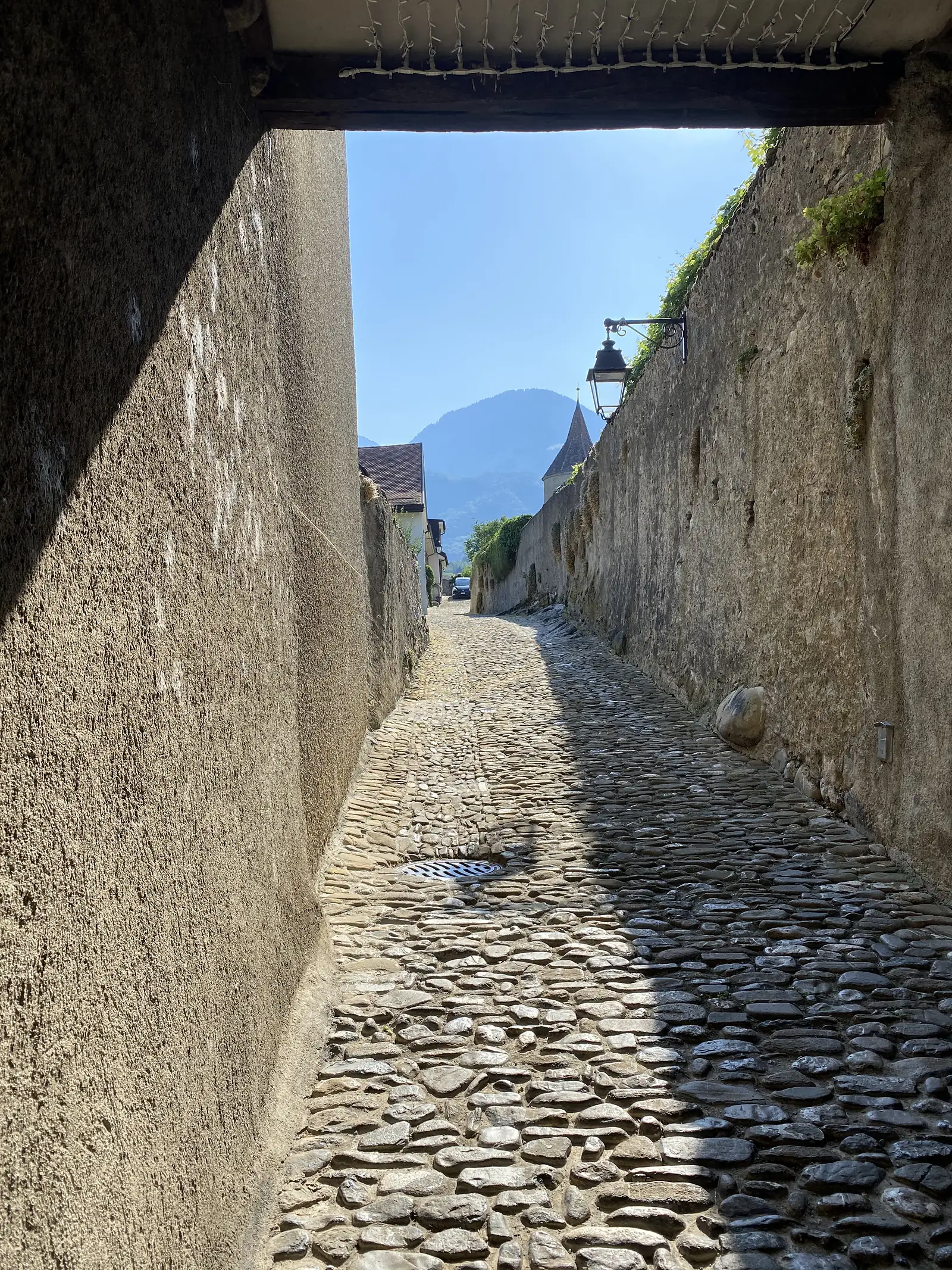 A cobbled lane runs beneath a stone gateway between old town walls, a lantern and a distant turret visible where the passage opens onto a village street.