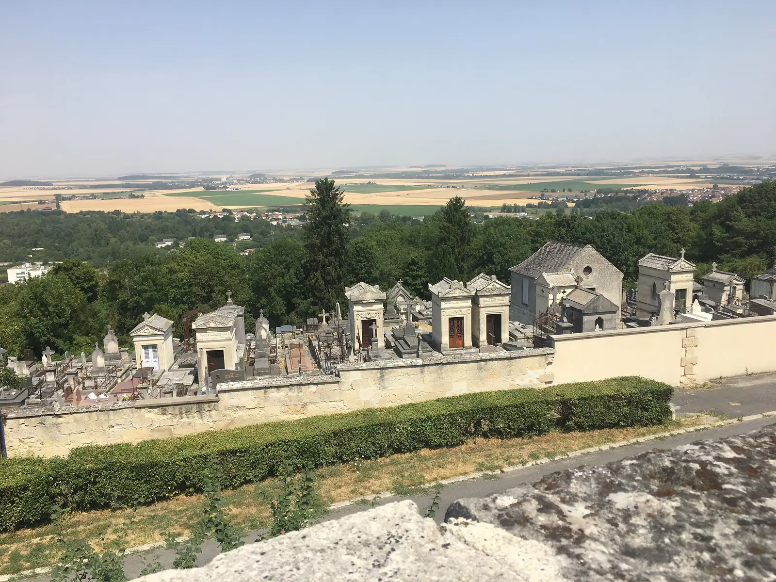 Rows of stone family tombs and crosses fill a hilltop cemetery, with a wooded slope and a wide patchwork of farmland and rooftops spreading out below.