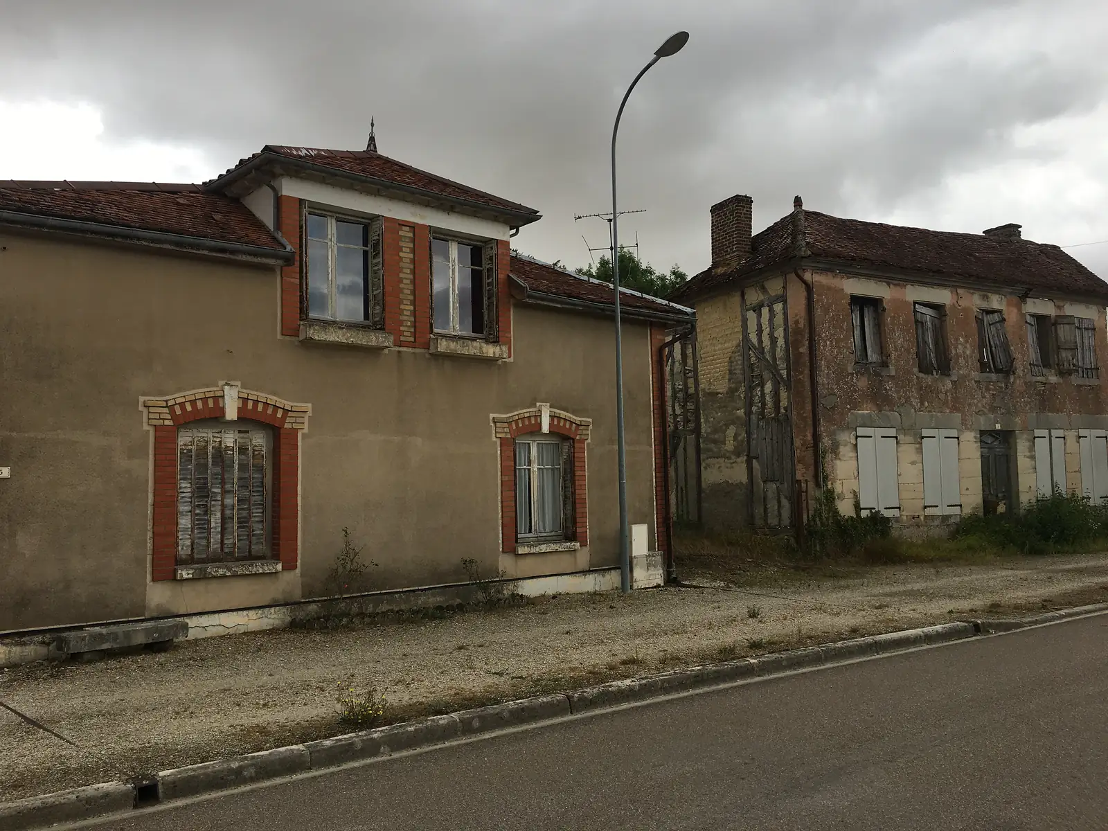 Two neglected stone houses with shuttered windows and a peeling chimney stand empty along a street beneath a heavy sky.