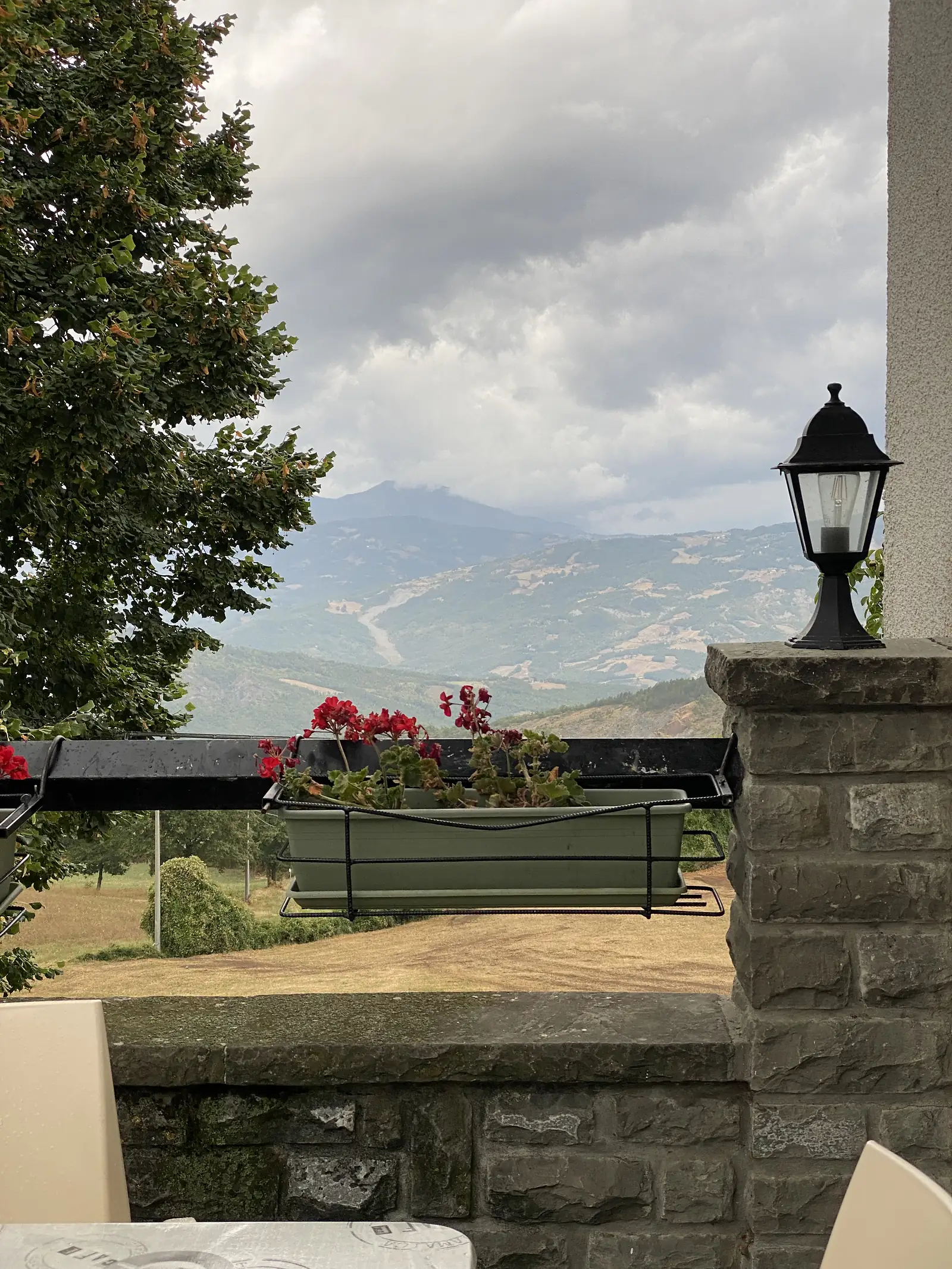 A wrought-iron lamp post and a window box of red geraniums frame a hazy view over layered mountain ridges from a stone terrace.