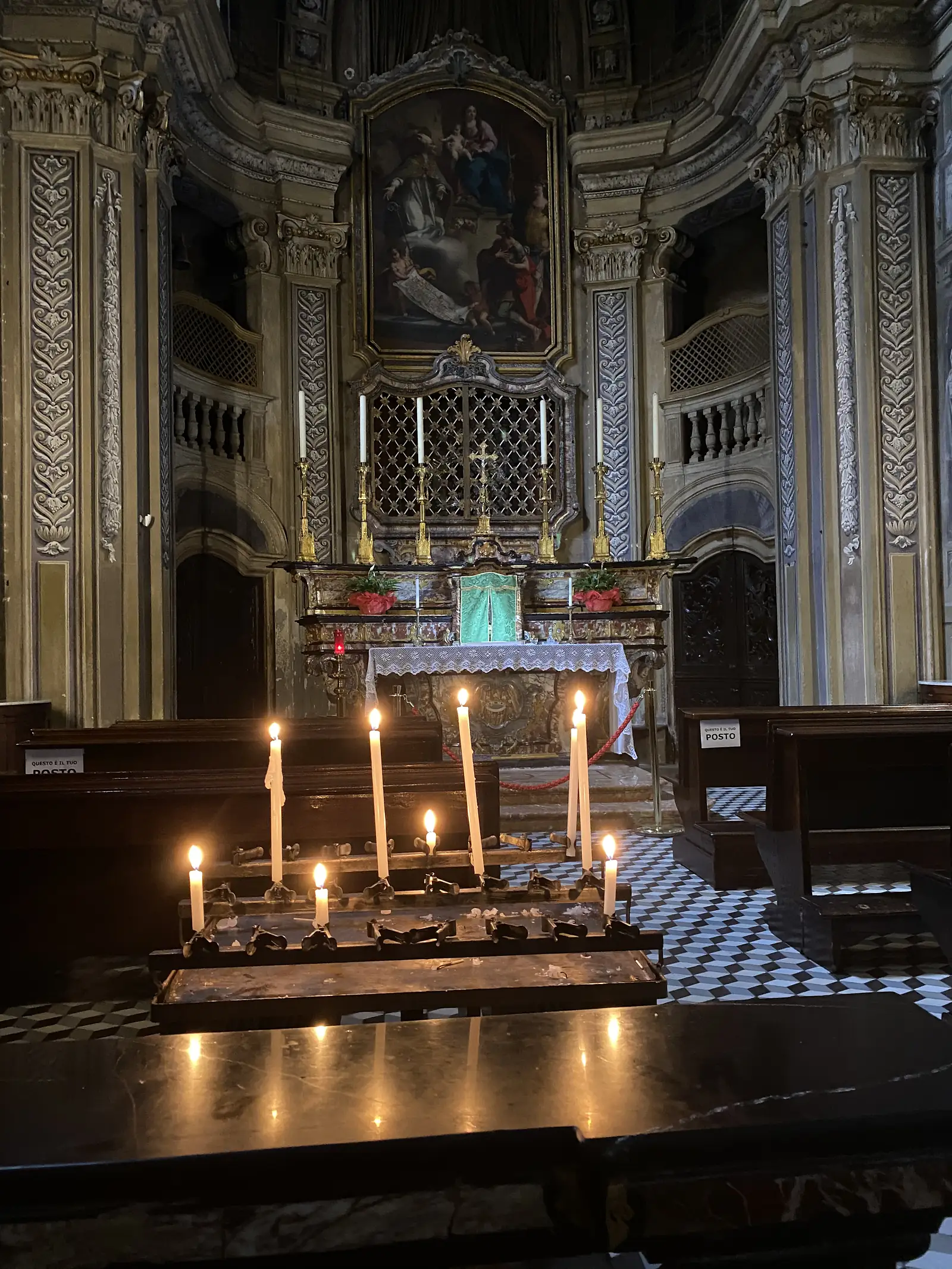 A row of lit votive candles in the foreground of a Baroque church interior, with a green-draped tabernacle, gilded candlesticks and a framed religious painting on the altar beyond.