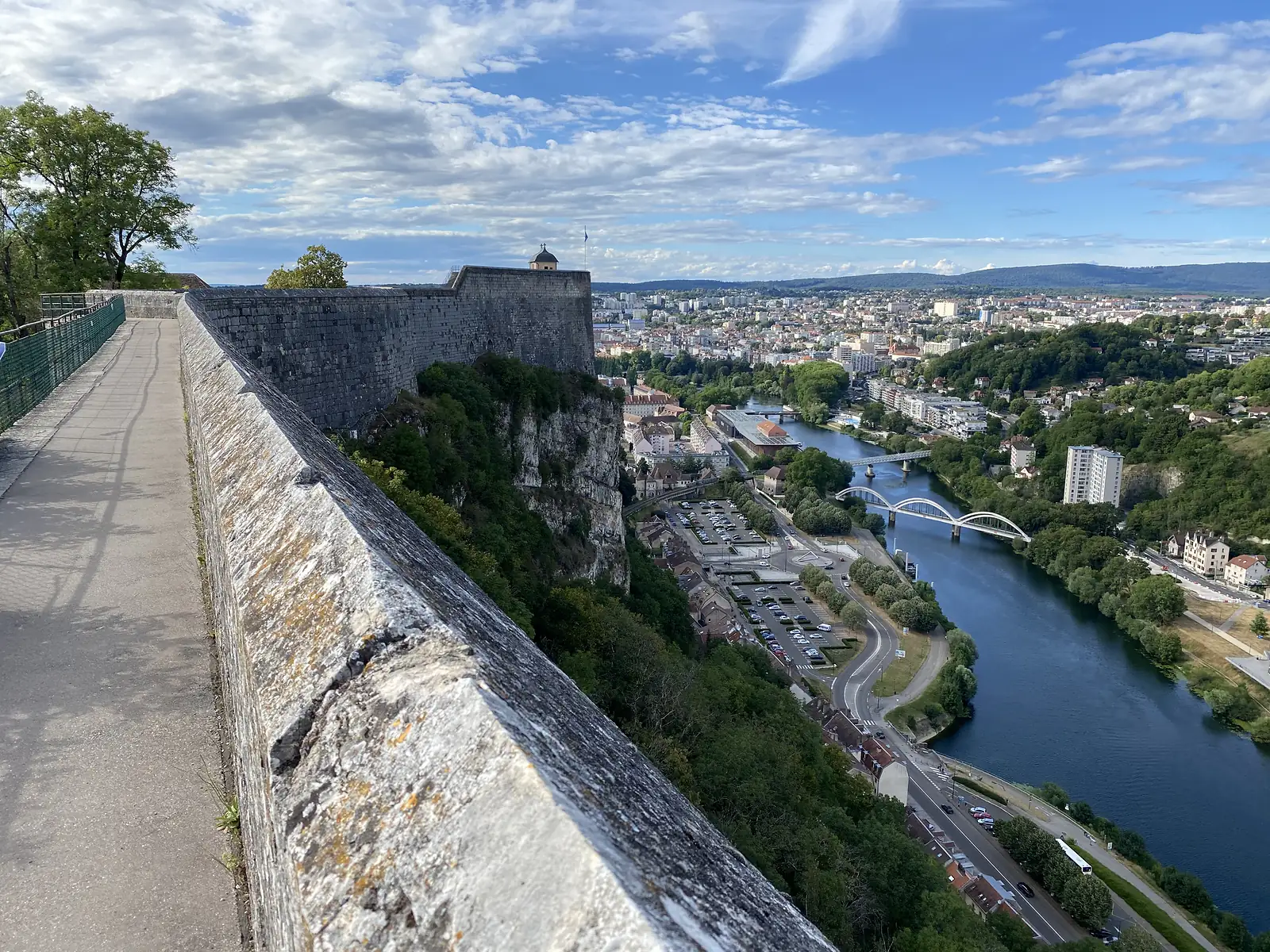 The Doubs river curves past a bridge and rooftops below a citadel wall, with wooded hills rising beyond the town.