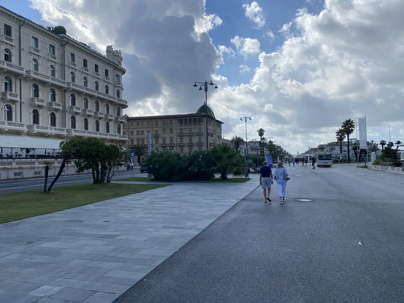 A couple walk along a broad paved seafront promenade past an ornate cream hotel building with balconied façades, palm trees and a bus stopped further along the road.